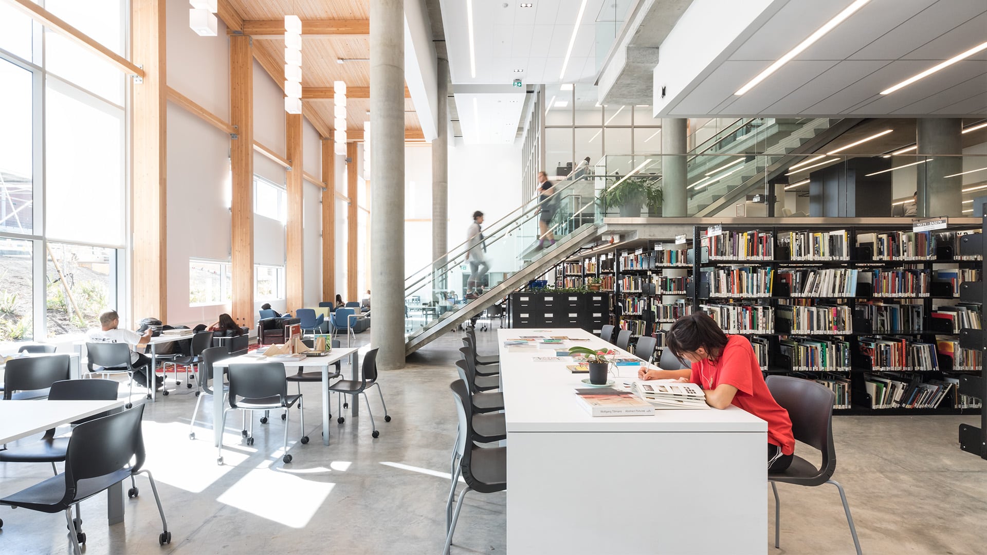A spacious and modern library at Emily Carr University of Art + Design, featuring high ceilings with exposed wooden beams, large windows allowing natural light to flood in, and rows of bookshelves filled with books. Students are seated at tables, studying and working, while others walk up a staircase in the background. The environment is bright, open, and conducive to learning.