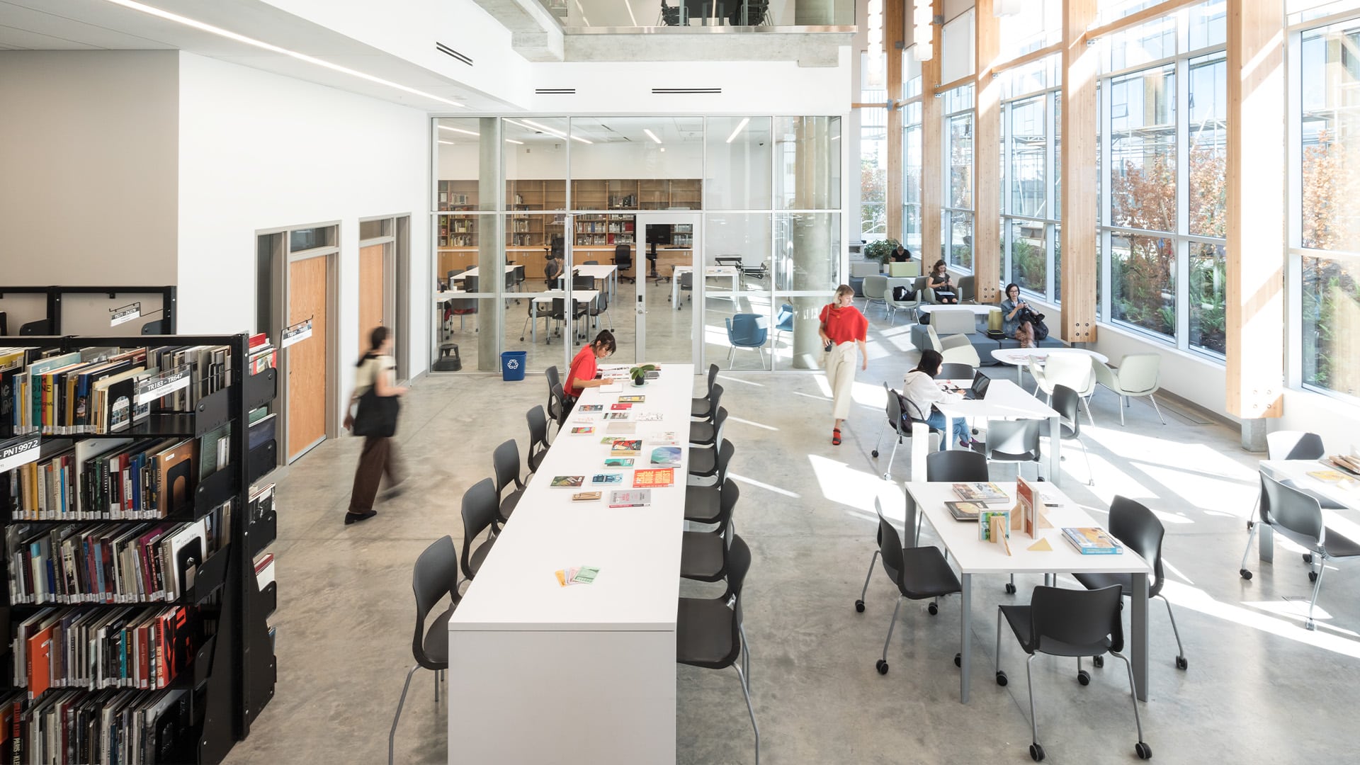 A bright and modern library space at Emily Carr University of Art + Design, featuring large windows that let in ample natural light. The area includes rows of bookshelves filled with books, long study tables with chairs, and comfortable seating areas. Students are seen walking, studying, and working on their laptops, creating a dynamic and collaborative atmosphere. The design incorporates wooden beams and open spaces, enhancing the sense of openness and creativity.