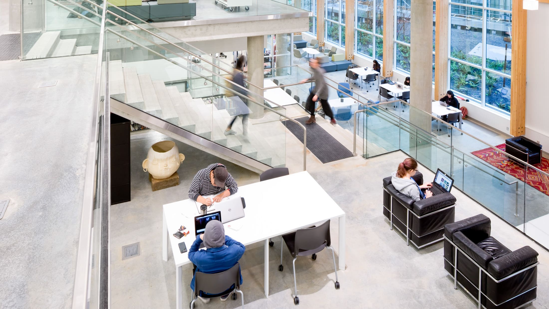 A spacious and modern library or study area at Emily Carr University of Art + Design, featuring an open layout with multiple levels. Students are seated at tables and chairs, working on laptops and engaging in collaborative activities. The space includes comfortable seating areas, large windows allowing natural light to flood in, and a staircase connecting different floors. A sculpture is visible near the center, adding an artistic touch to the environment.