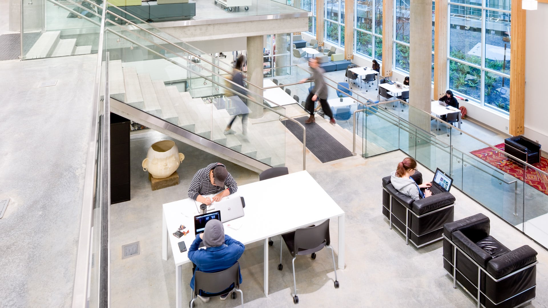 A spacious and modern library or study area at Emily Carr University of Art + Design, featuring an open layout with multiple levels. Students are seated at tables and chairs, working on laptops and engaging in collaborative activities. The space includes comfortable seating areas, large windows allowing natural light to flood in, and a staircase connecting different floors. A sculpture is visible near the centre, adding an artistic touch to the environment.