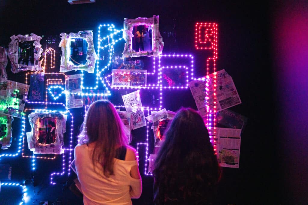 Two viewers observe a neon-lit wall installation of photos, newspaper clippings, and LED outlines at the 2025 Ghost Images – Photography and Trace exhibition.