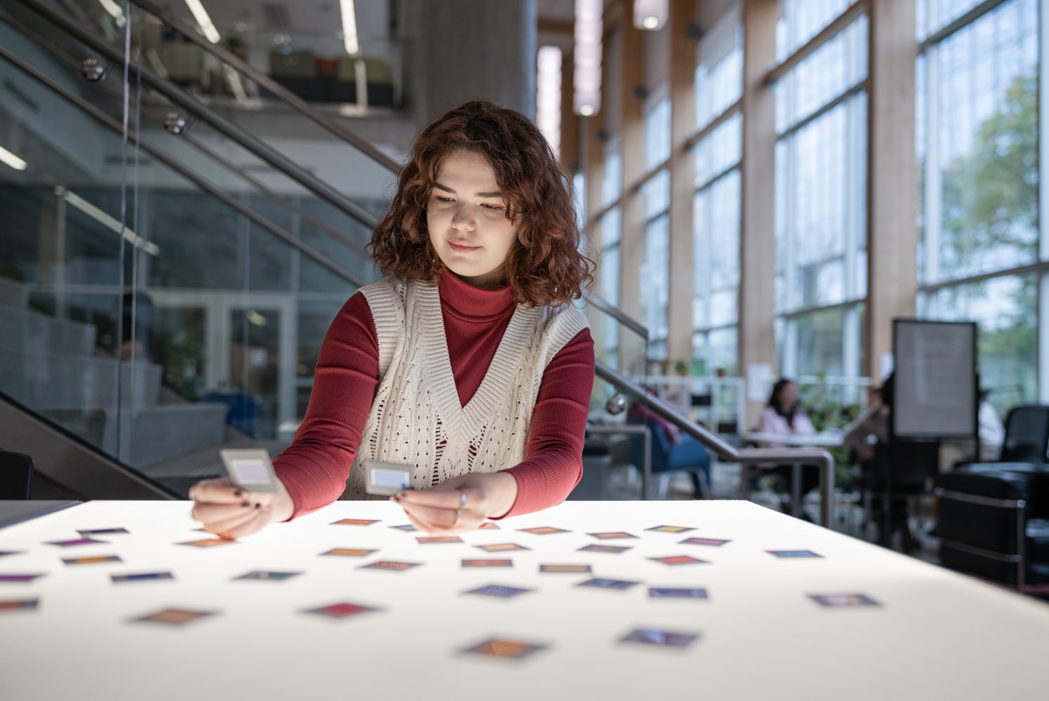 A woman is seated at a table in a modern, well-lit space, carefully arranging colorful cards or tiles on the surface. She appears focused and engaged, holding a small object in one hand while sorting through the items on the table. The background features large windows, allowing natural light to illuminate the area, and other individuals can be seen working in the distance.