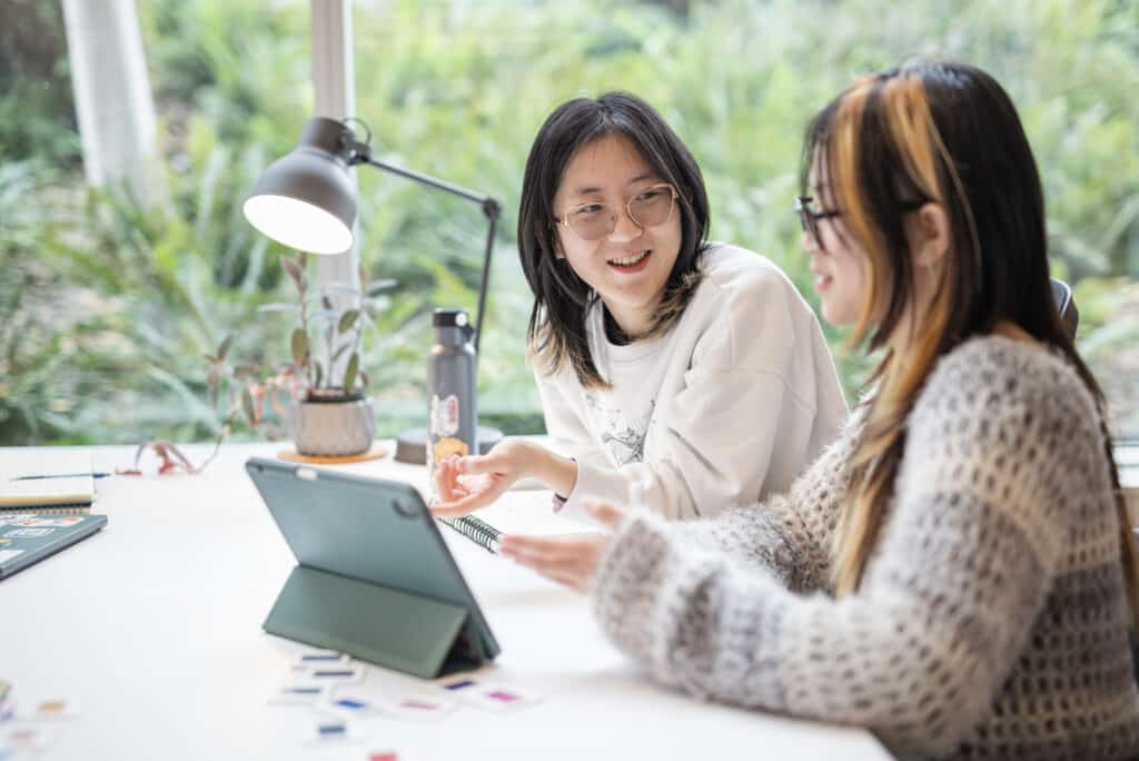 Two students are engaged in a collaborative discussion at a table, using a tablet and working with colorful cards or materials. The setting is bright and inviting, with natural light streaming in through a window that offers a view of greenery outside. One student gestures enthusiastically while explaining something, while the other listens attentively, both smiling and appearing focused on their task.