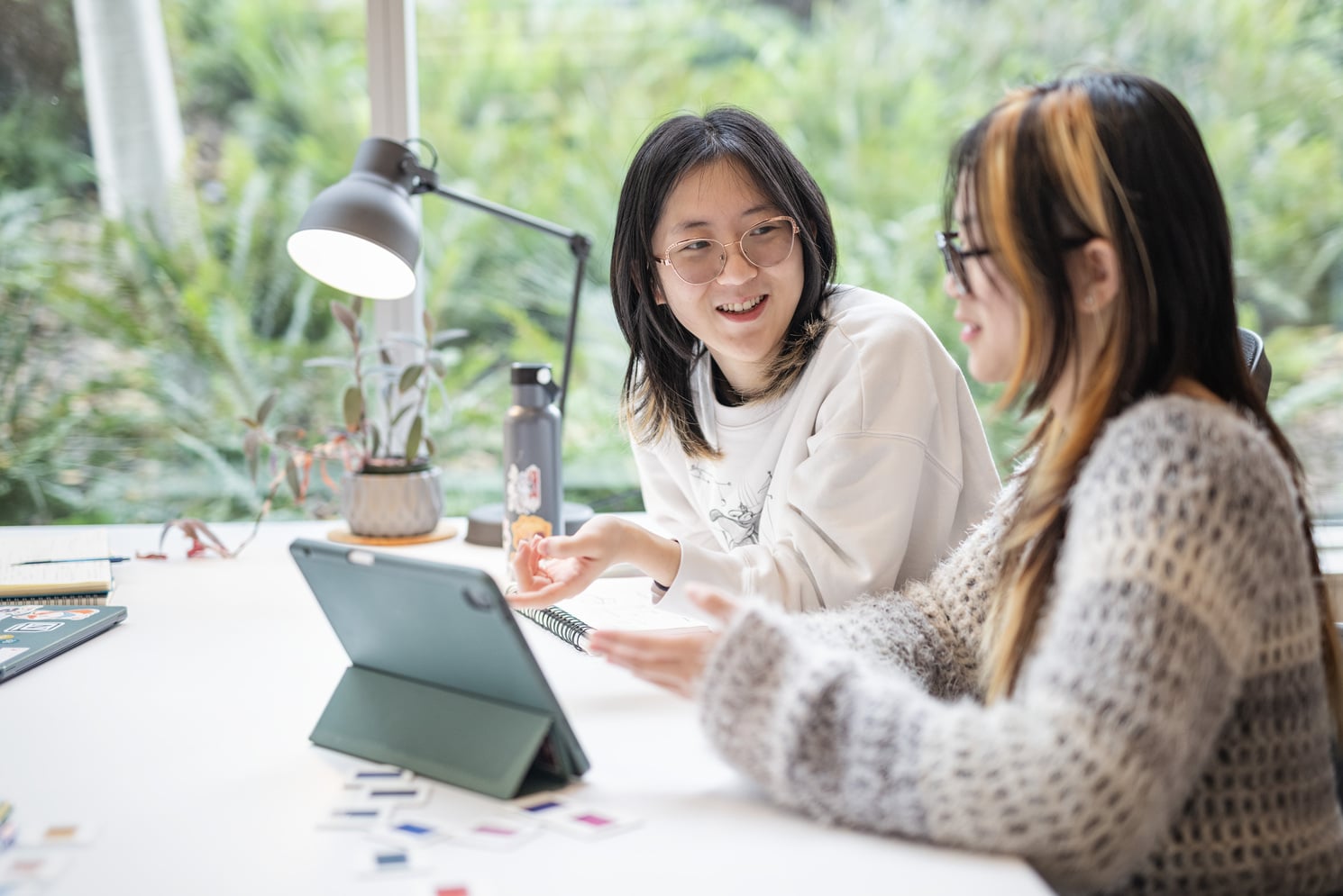 Two students are engaged in a collaborative discussion at a table, using a tablet and working with colorful cards or materials. The setting is bright and inviting, with natural light streaming in through a window that offers a view of greenery outside. One student gestures enthusiastically while explaining something, while the other listens attentively, both smiling and appearing focused on their task.