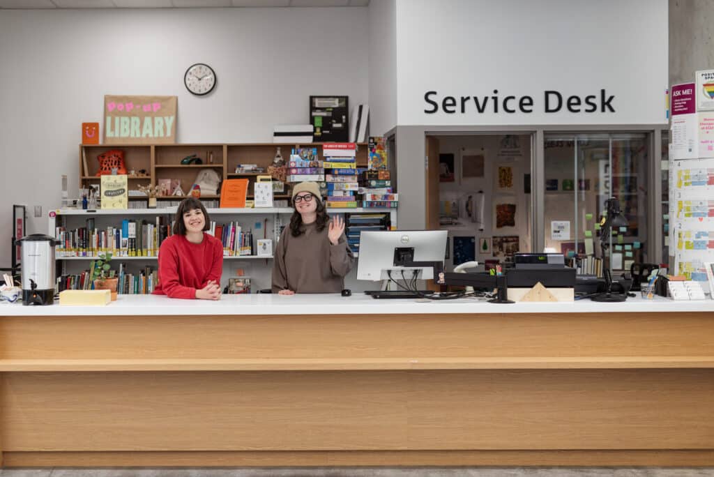 Two library staff members are standing behind a wooden service desk at the Emily Carr University of Art + Design Library. The person on the left is wearing a red sweater and smiling, while the person on the right is wearing a brown sweater and a beige beanie, waving hello. Behind them, shelves are filled with books, colorful posters, and decorative items. A sign above the desk reads 'Service Desk,' and there is a clock on the wall in the background.