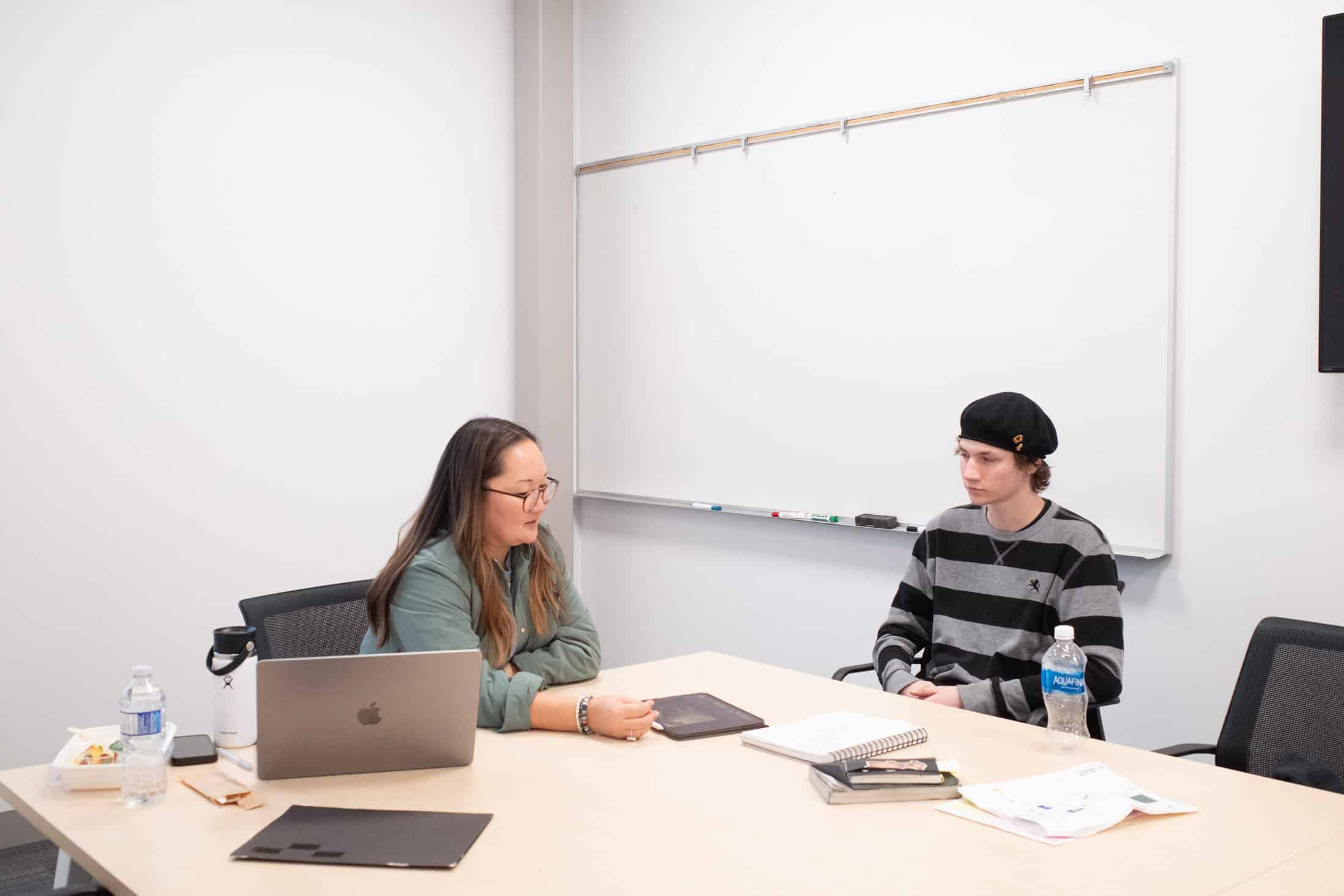 Two individuals are seated across from each other at a table in a meeting or study room. The person on the left, wearing glasses and a green jacket, is focused on a laptop, while the person on the right, wearing a striped sweater and a black beanie, looks attentively toward the other individual. The table has various items, including water bottles, notebooks, and papers, suggesting a collaborative or instructional setting. A whiteboard is visible in the background.