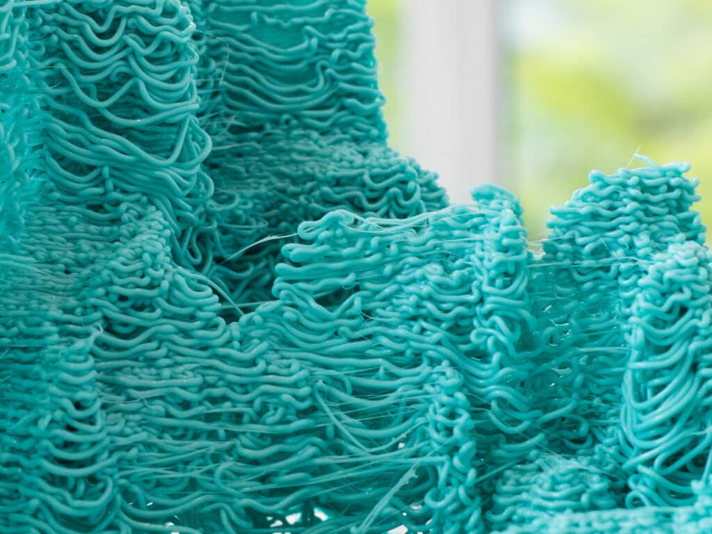 A close-up view of the art piece titled "What's Mine Is Yours" by Benjamin Mignault, BDes Industrial Design. The artwork features intricate digital embroidery on found fabric, showcasing a vibrant turquoise color with detailed, wavy patterns created through layered stitching. The texture is enhanced by 3D printing and laser etching techniques, resulting in a tactile and visually engaging surface. The interplay of traditional textile methods with modern technology highlights themes of craftsmanship, innovation, and the fusion of old and new.