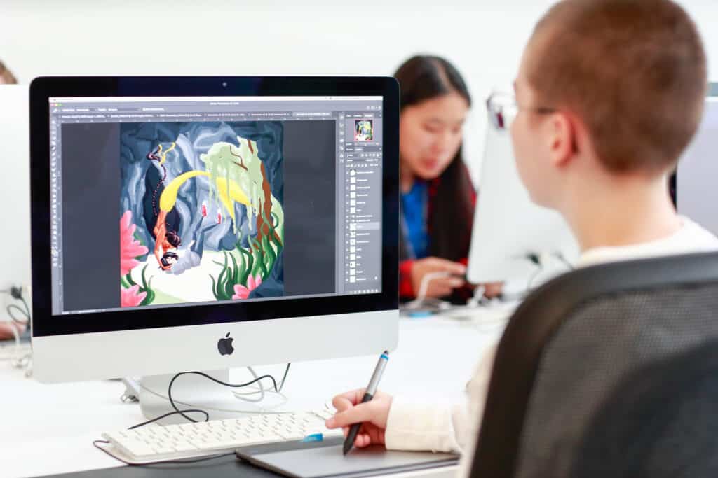 A student is using a Mac computer in the Emily Carr University Library to work on digital art, with Adobe Photoshop open on the screen displaying a colourful illustration. The workspace is well-lit, and another individual can be seen working at a nearby computer in the background.