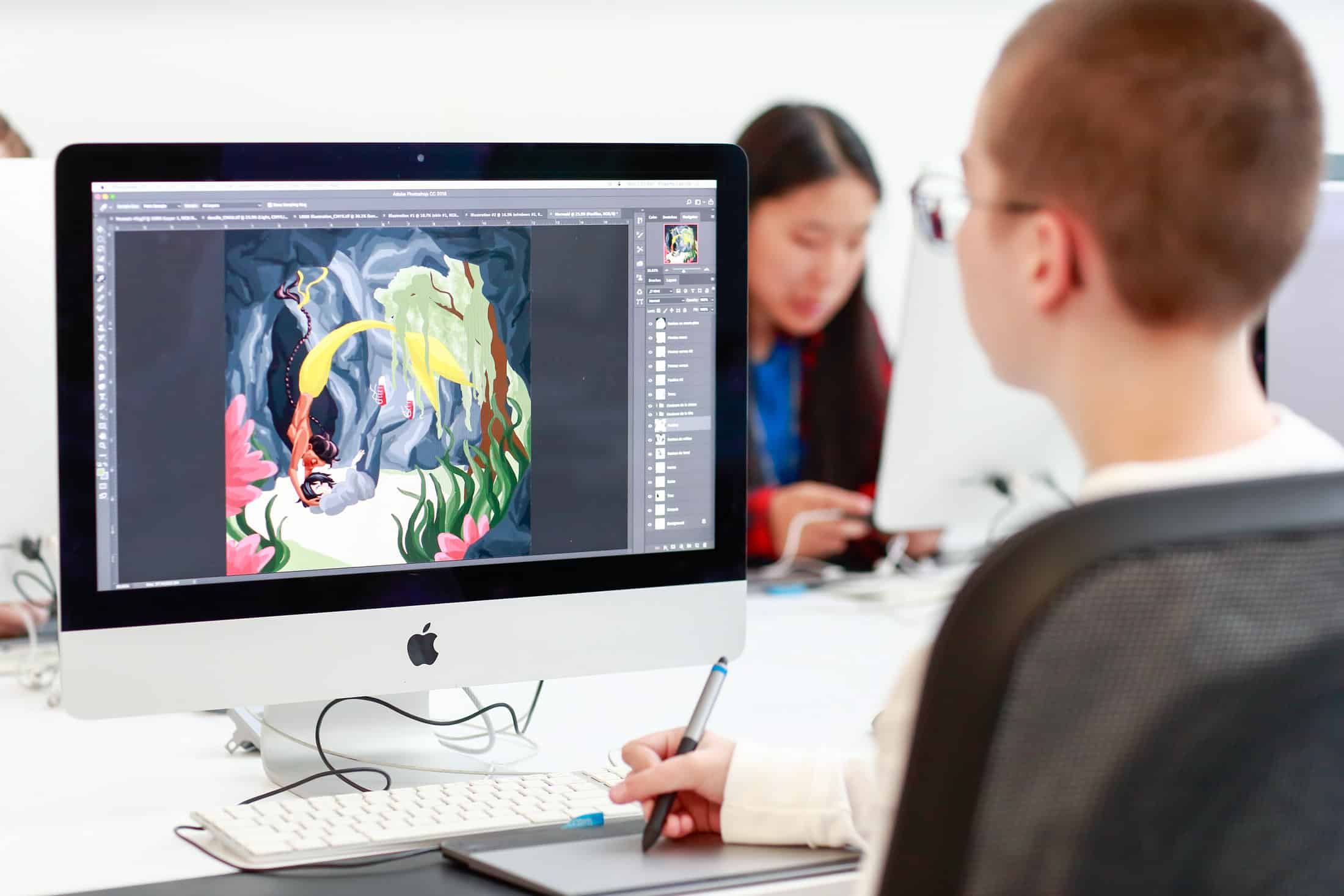 A student is using a Mac computer in the Emily Carr University Library to work on digital art, with Adobe Photoshop open on the screen displaying a colourful illustration. The workspace is well-lit, and another individual can be seen working at a nearby computer in the background.