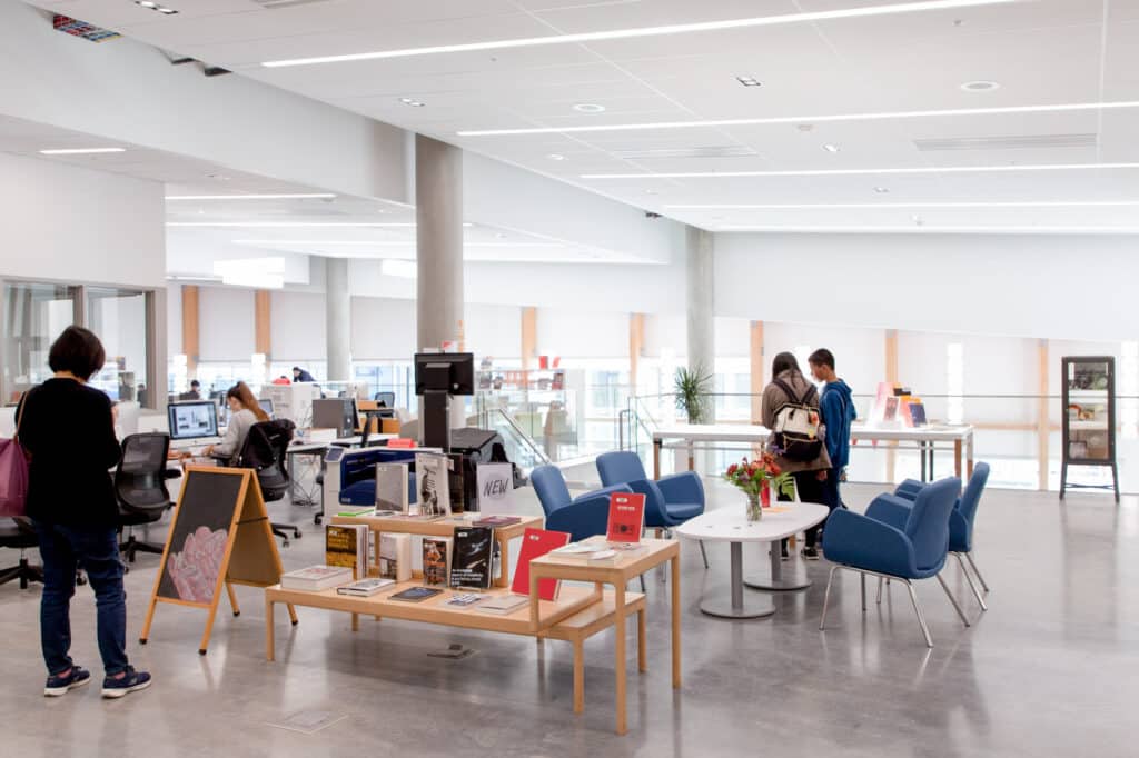 A modern library space with a bright and open design, featuring comfortable seating areas and workstations. Students are engaged in various activities, such as working on computers and browsing materials. The area includes tables with books and magazines, a welcome sign, and large windows that allow natural light to fill the room. The environment is conducive to both individual study and collaborative learning.
