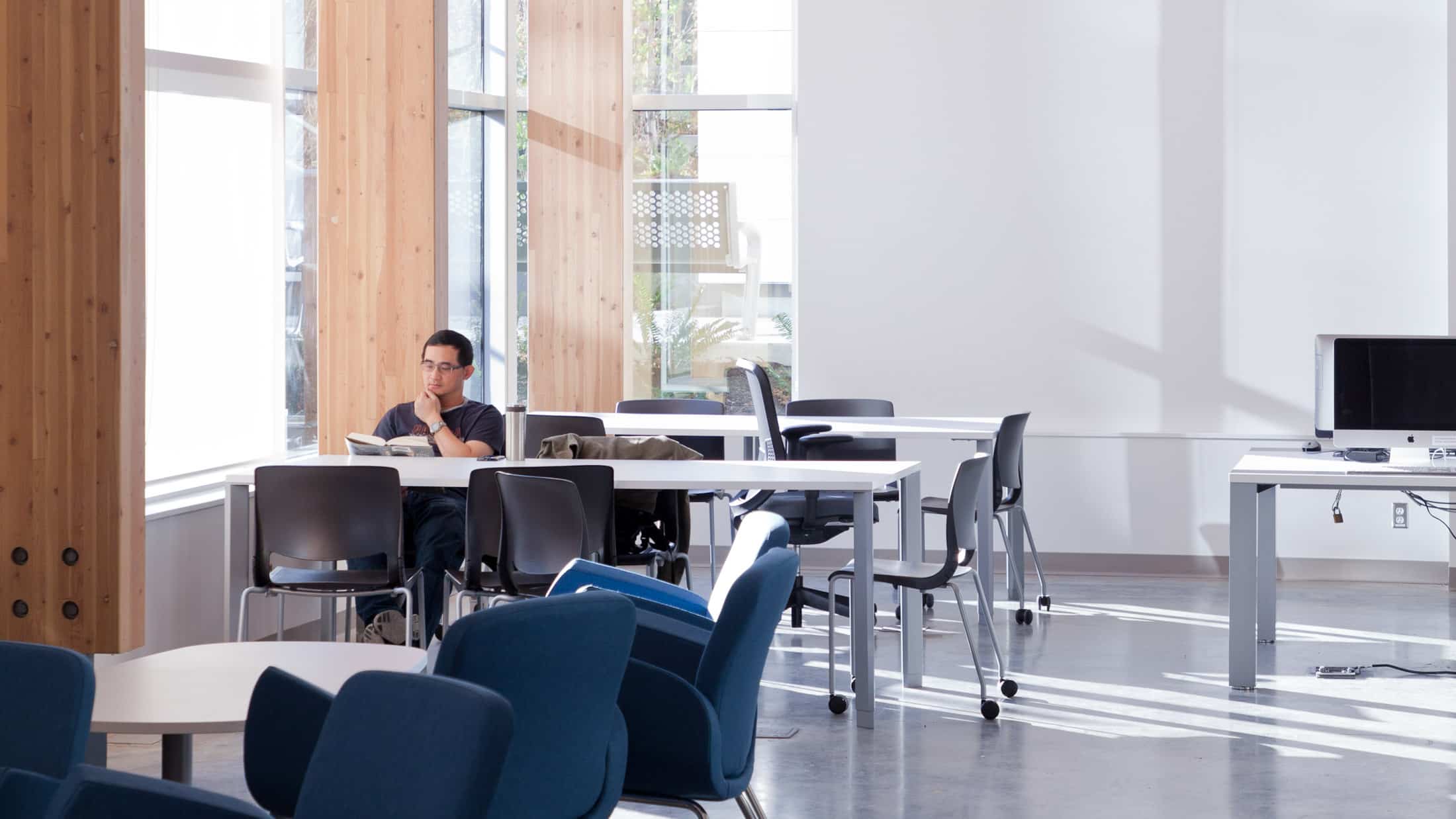 A student is seated at a table in a bright, modern study area, reading a book. The space features large windows that allow natural light to flood in, creating a calm and focused environment. The room includes multiple tables and chairs, some of which are occupied by other students, while others remain empty. Wooden beams and white walls contribute to the clean and organized aesthetic