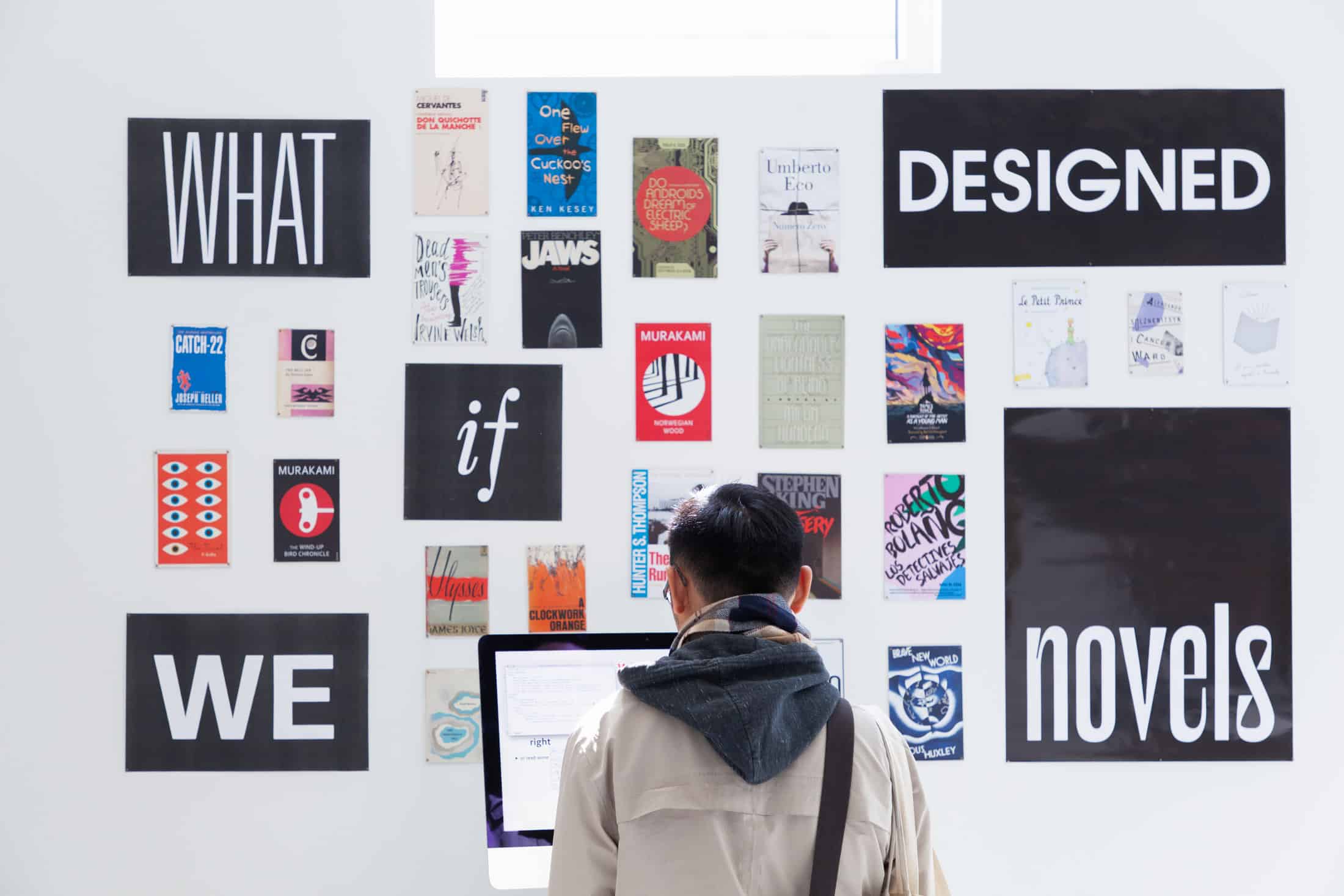 A man stands in front of an exhibition wall displaying a curated collection of book covers and posters. The wall features bold text reading 'WHAT if WE DESIGNED novels,' encouraging viewers to reflect on the intersection of design and literature. The arrangement includes various artistic elements, such as colorful book covers and abstract designs, fostering inspiration and creative thinking.