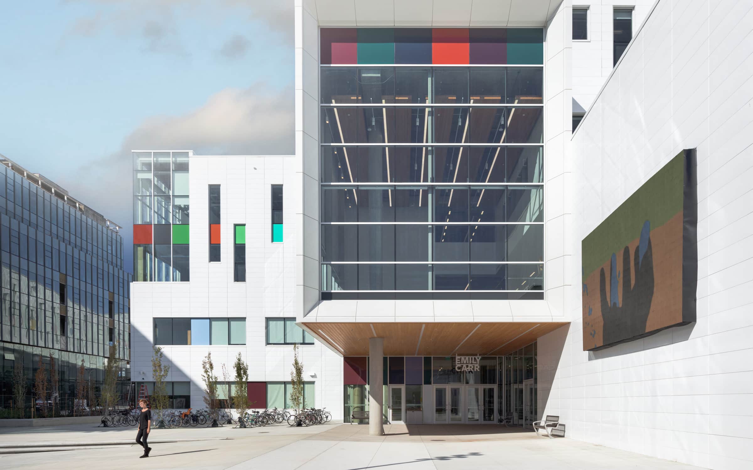 The Urban Screen and East Entrance at Emily Carr University showcase a modern architectural design with vibrant colors and large glass windows. The building features a prominent digital display screen on the side, adding a dynamic visual element to the campus. The entrance is marked by wooden accents and clear signage, creating an inviting and contemporary atmosphere.
