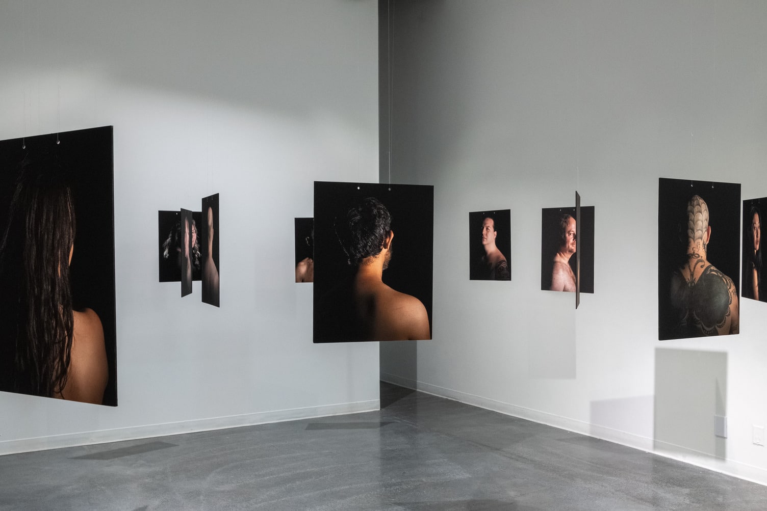 Photographic exhibition showing large portraits of people’s backs and profiles, printed on black backgrounds and suspended from the ceiling in a gallery space.