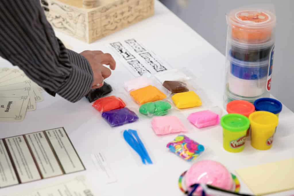 A vibrant table setup at the Health Design Lab's 2024 Showcase at Emily Carr University features a variety of colorful materials, including playdough in bright hues like orange, yellow, green, and pink. The table also includes containers of markers, buttons, and other creative tools, inviting participants to engage in hands-on activities. A hand reaches for one of the playdough packets, highlighting the interactive and collaborative nature of the event.