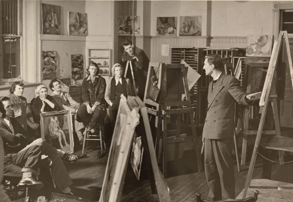 A black-and-white photograph of an art classroom, likely from the mid-20th century. A male instructor stands beside an easel, addressing a group of students seated around him. The students, dressed in mid-century clothing, listen attentively, with paintings and canvases visible throughout the room and mounted on the walls behind them.