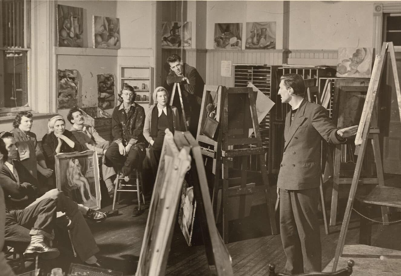 A black-and-white photograph of an art classroom, likely from the mid-20th century. A male instructor stands beside an easel, addressing a group of students seated around him. The students, dressed in mid-century clothing, listen attentively, with paintings and canvases visible throughout the room and mounted on the walls behind them.