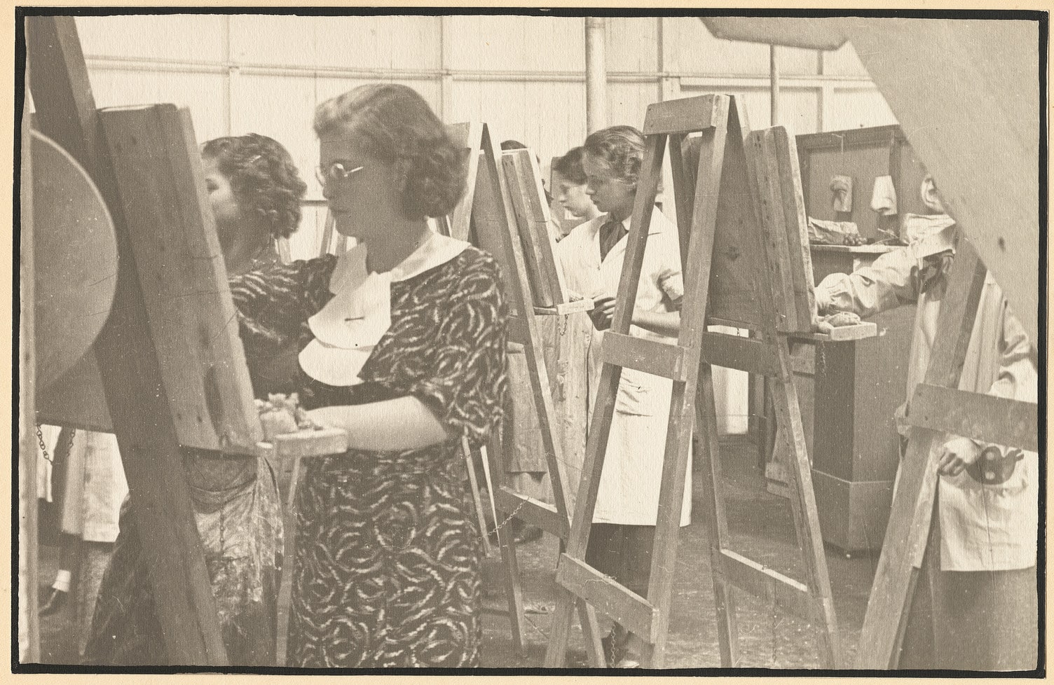 A sepia-toned photograph of an art classroom showing several women painting at wooden easels. The students, wearing dresses or smocks, concentrate on their canvases in a well-lit studio with high walls and plaster casts visible in the background.