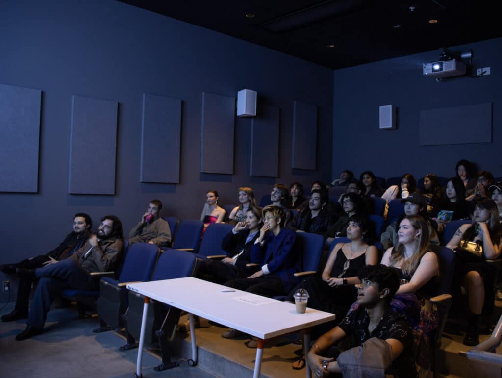 A group of attendees is seated in a darkened theater at Emily Carr University of Art + Design, watching dynamic media screenings during the 2025 show. The audience is focused and engaged, with some holding drinks or snacks, while others lean forward to better absorb the content. The room features soundproofing panels on the walls and a projector screen displaying vibrant visuals, creating an immersive viewing experience.