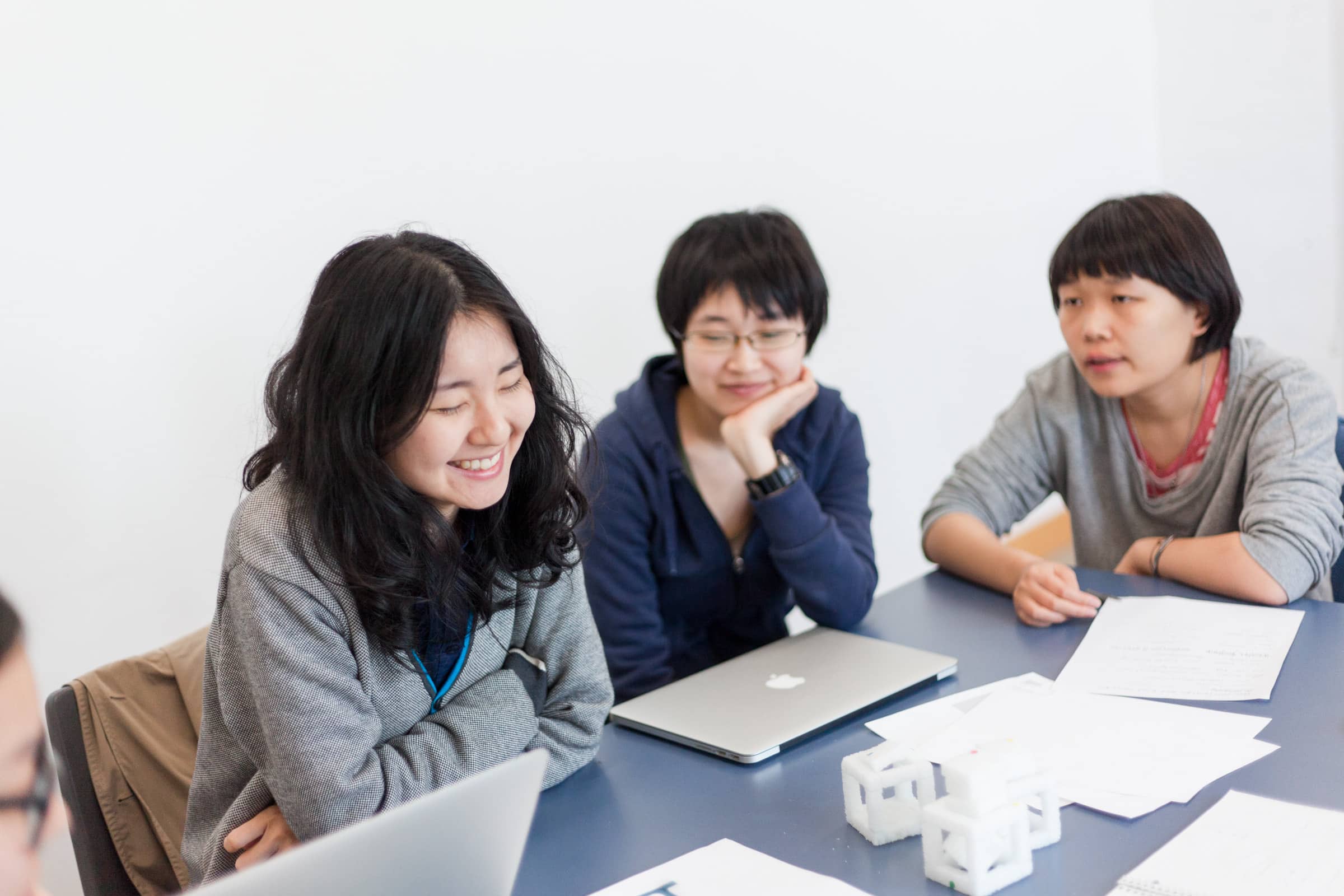 Master of Design students collaborate in a lively and engaging session, seated around a table equipped with laptops and printed materials. The atmosphere is dynamic, with one student smiling broadly, indicating a moment of shared laughter or enjoyment during their discussion. The setting reflects a focused yet positive environment, emphasizing teamwork and creative problem-solving.