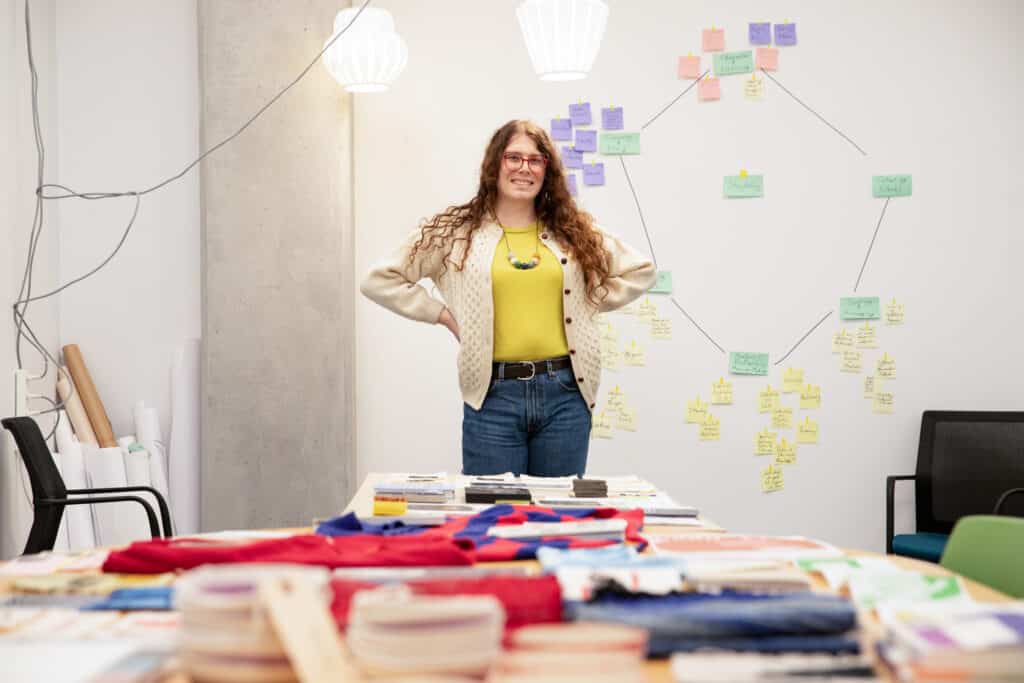 Master of Design student Morgan Martino stands confidently in the DESIS Lab at Emily Carr University during the Jake Kerr Faculty of Graduate Studies 2024 Open Studios. The background features a wall covered with colorful sticky notes and diagrams, showcasing her research and design process. The table in front of her is organized with neatly folded fabrics and materials, reflecting her creative and methodical approach to her work.