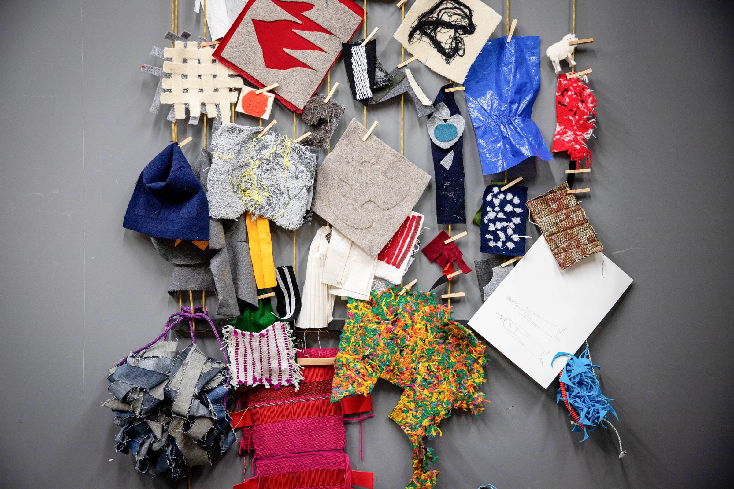 The Material Matters research and production lab at Emily Carr University of Art + Design showcases a vibrant display of textile and fabric samples. Various materials, including felted pieces, woven fabrics, and handcrafted items, are hung on strings against a dark background. The collection features a mix of bold colors, intricate patterns, and textures, highlighting the creativity and experimentation that takes place in this innovative space.