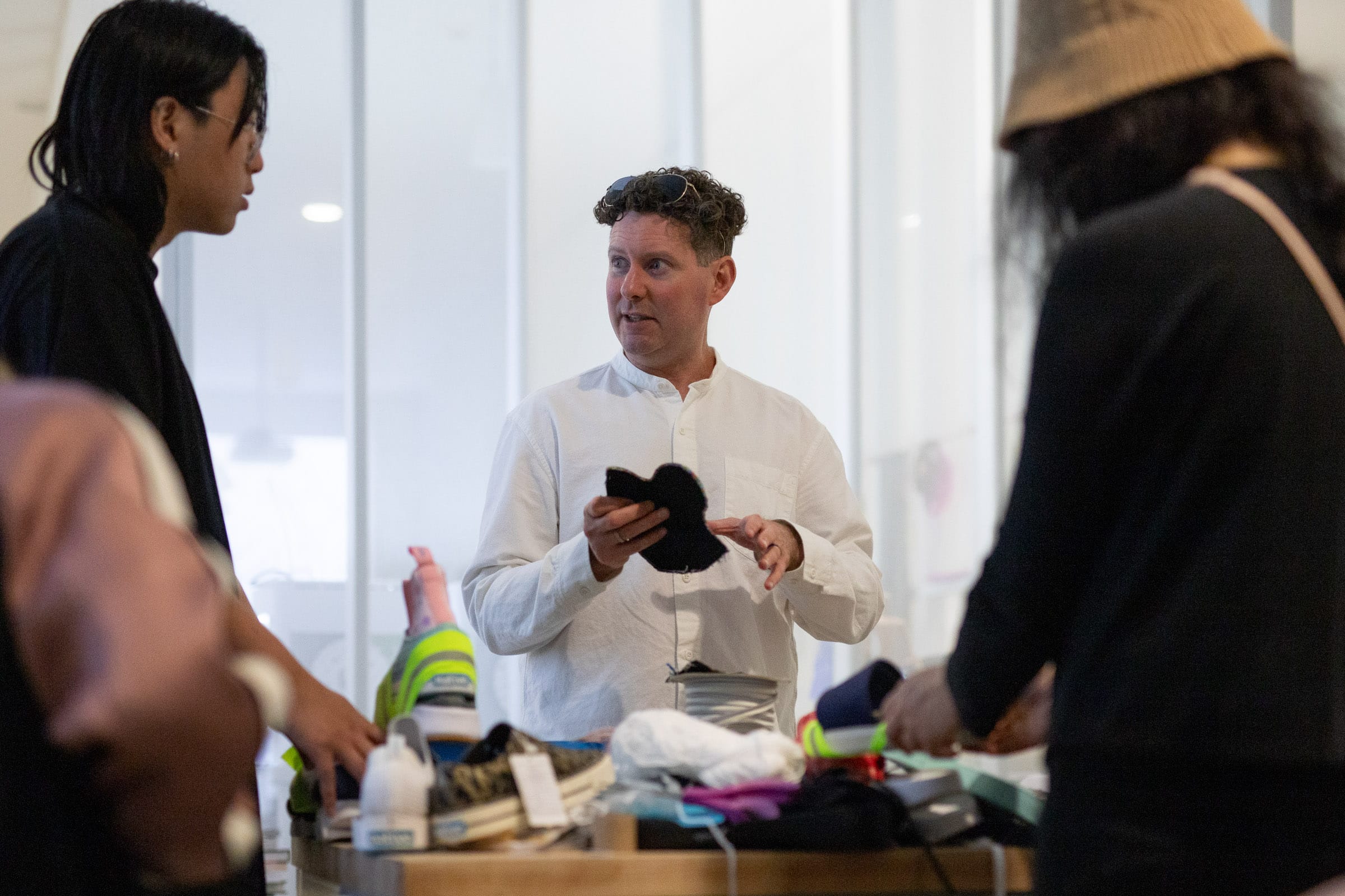 During the Native Shoe Hack event at the Material Matters satellite lab at the Vancouver Art Gallery, ECU students collaborate with staff, faculty members Hélène Day Fraser and Keith Doyle, and Native Shoes co-founder Tom Claypool. In this image, participants are engaged in a discussion while working on deconstructing and reassembling existing Native shoes. The table is filled with various materials and tools, highlighting the hands-on nature of the project.