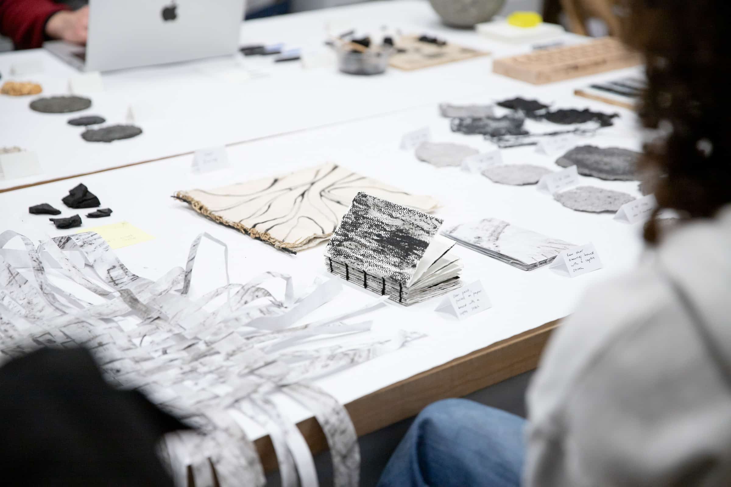 A table is filled with a variety of early iterative materials created as part of the Charred Wood research initiative at Emily Carr University. The display includes textured fabrics, woven pieces, and other experimental materials made from wood charcoal. Each item is labeled, showcasing the diverse forms and applications being explored by researchers and students. The setting reflects a collaborative and creative environment focused on sustainable design.