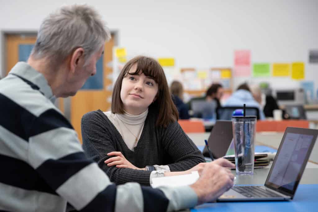 A student engages in a focused discussion with an instructor or mentor during a workshop or collaborative session. The setting is a bright, modern classroom or studio environment, characterized by colorful sticky notes on the wall and other participants working in the background. The student appears attentive and thoughtful, while the mentor gestures toward a laptop screen, suggesting a hands-on learning experience centered around technology and creative problem-solving.