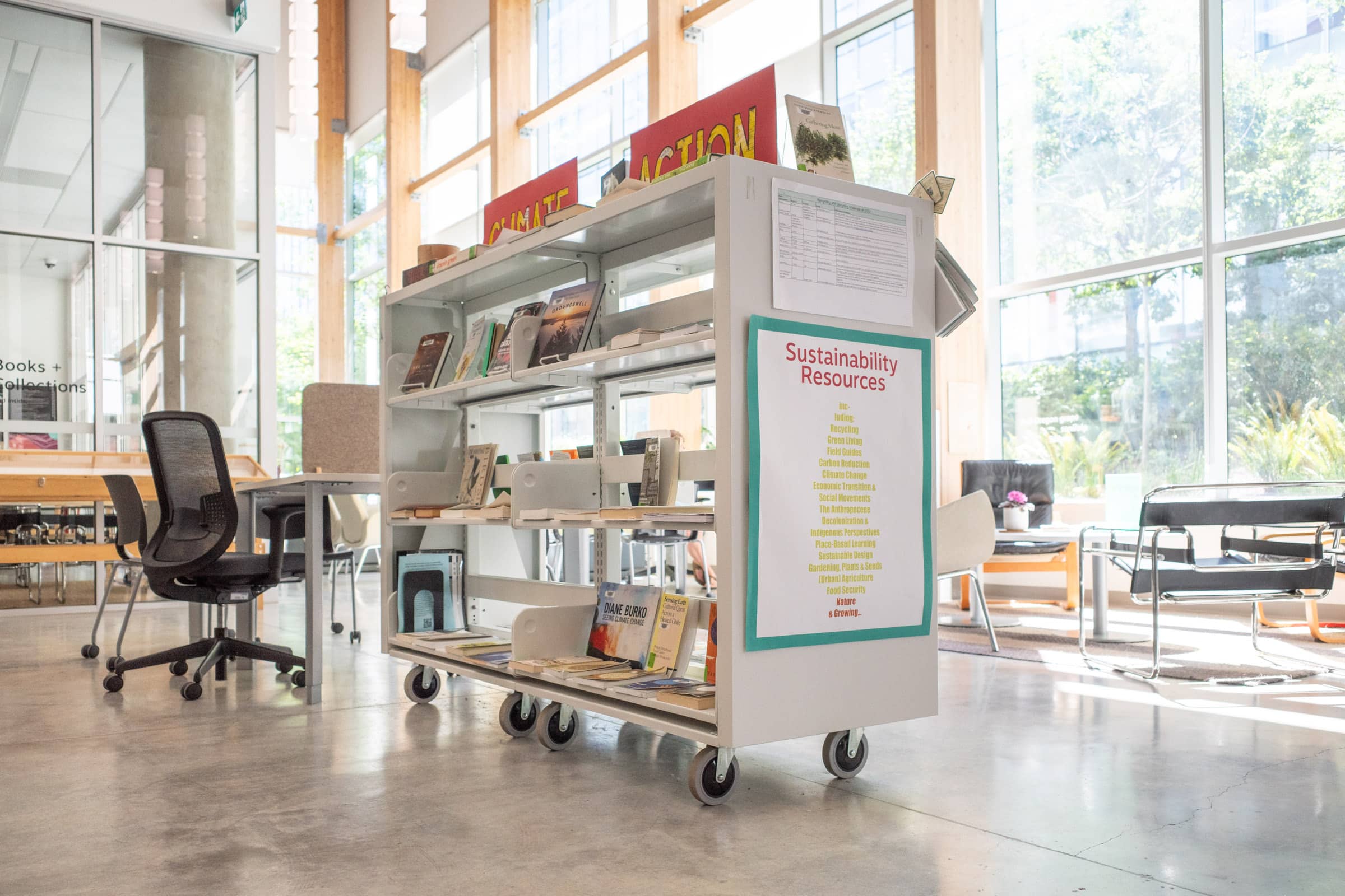 A mobile cart in the Emily Carr University Library is dedicated to sustainability resources. The cart features shelves filled with books and materials on topics such as recycling, green living, climate change, and more. A sign on the side lists key themes like carbon reduction, economic transition, and sustainable design. The setting is bright and modern, with ample natural light streaming through large windows.