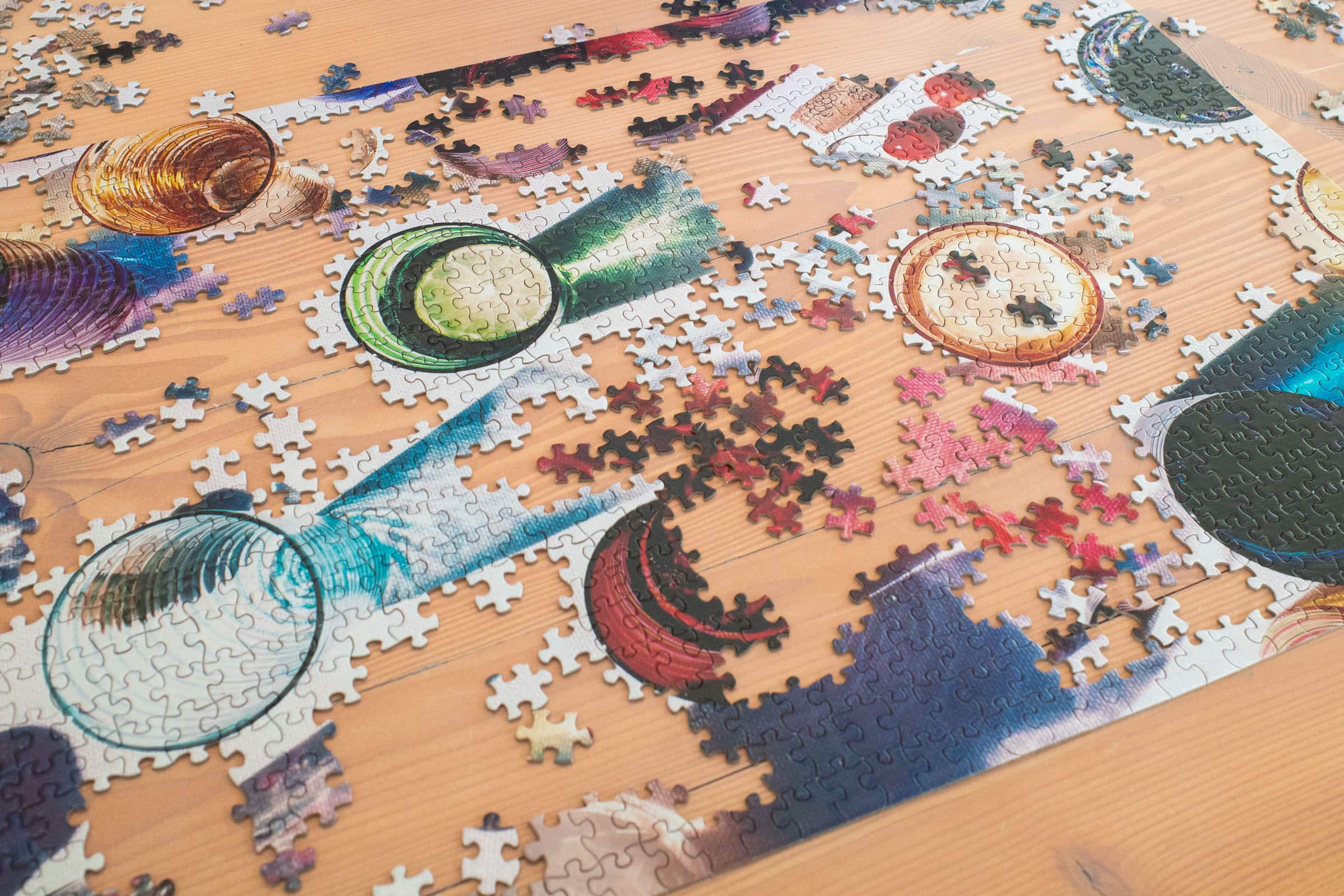 A colourful jigsaw puzzle is being assembled on a wooden table. The puzzle features vibrant and intricate details, including elements like seashells, a telescope, and celestial imagery. Several sections of the puzzle are completed, while others remain scattered around, indicating an ongoing activity. The scene suggests a relaxing and engaging pastime.