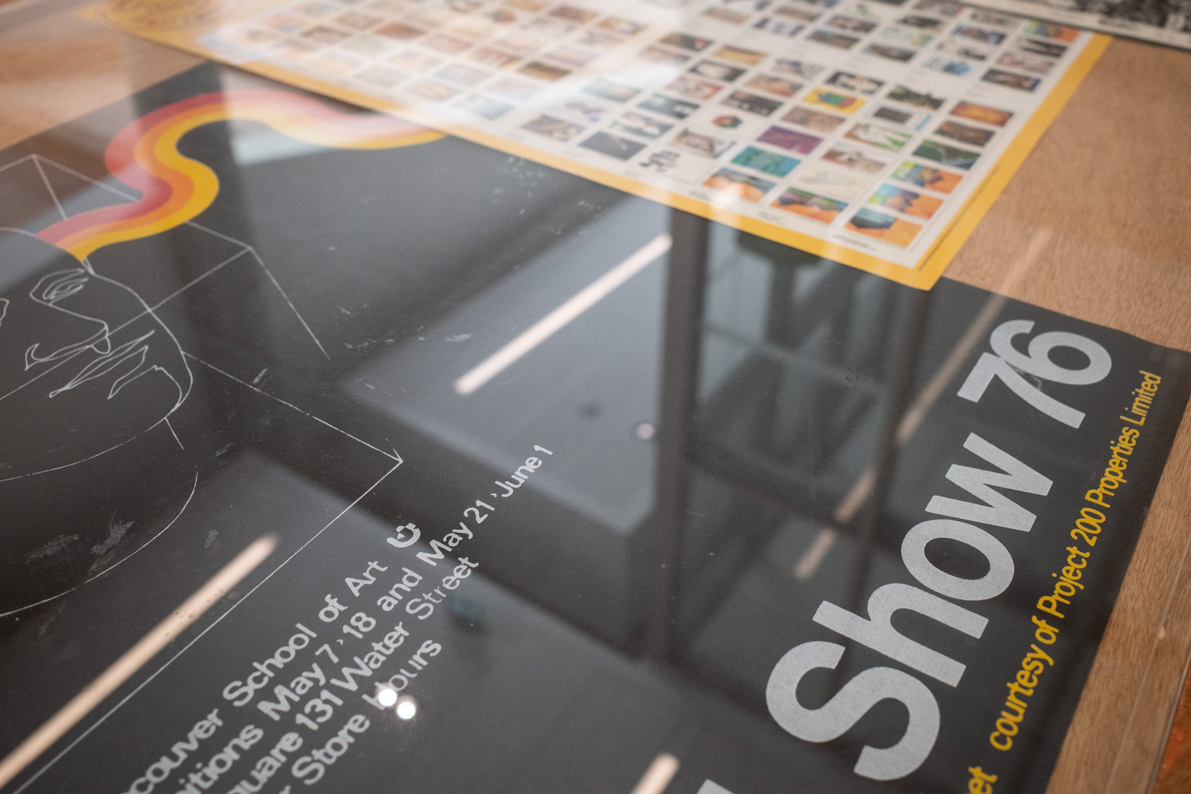 A display case in the Emily Carr University showcases a curated exhibition of creative works. The transparent acrylic case protects items such as colorful posters, sketches, and printed materials arranged neatly on a wooden surface. The exhibit includes abstract designs, text-based artwork, and a book cover featuring a vibrant image. The setup highlights the diversity of artistic expressions within the ECU community.