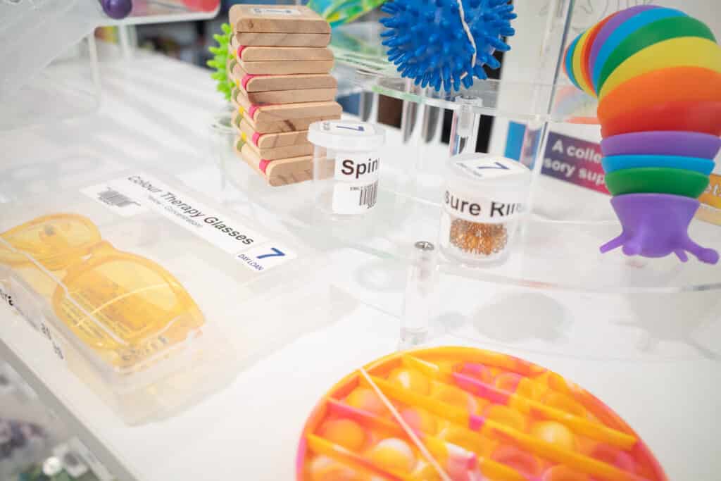 A display of sensory and neurotools items on a clear glass table in the ECU Library. The collection includes colorful objects such as a stack of wooden blocks, a blue textured ball, a set of rainbow-colored stacking rings, a fidget toy with popping bubbles, and labeled containers for items like 'Spin' and 'Sure Ring.' These tools are designed to support focus, relaxation, and sensory engagement.