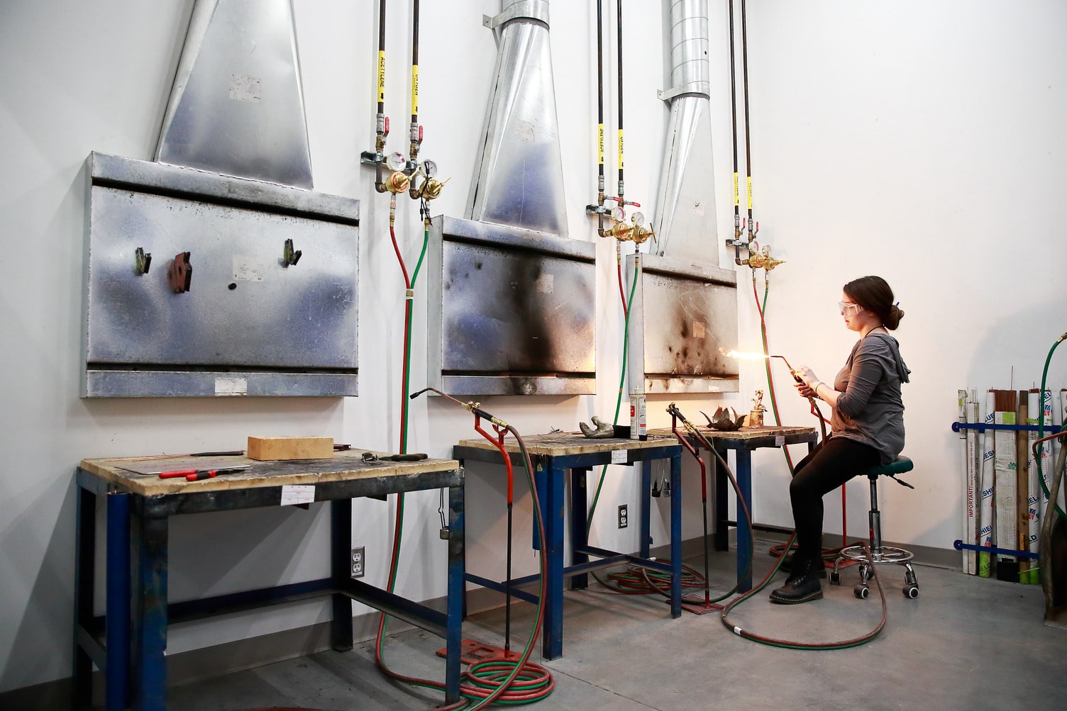 A student wearing safety goggles sits on a stool in a metalworking studio, using a handheld torch that emits a bright flame. She is focused on a small object on her workbench. Behind her are three large, industrial kilns or furnaces mounted on the wall, each with gas hoses and regulators. The room has white walls and concrete floors, with red and green hoses coiled on the ground and tools arranged on blue-framed workbenches.