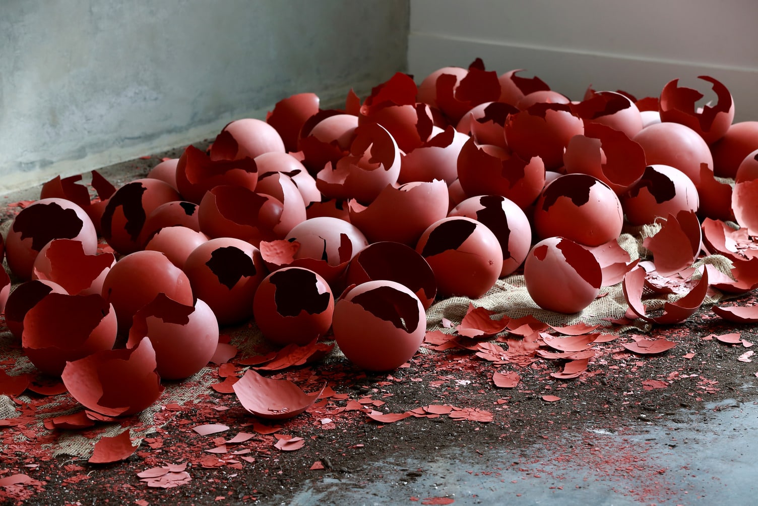 A pile of broken, matte red eggshells scattered across a rough, dark concrete floor. Many shells are cracked open or shattered into jagged pieces, revealing their hollow interiors. The fragments vary in size, with some larger halves and many smaller shards littering the ground. A coarse, light-colored fabric, possibly burlap, lies partially beneath the shells. In the background, a plain, textured wall meets the floor at a corner, creating a stark, minimalist setting that emphasizes the vivid red of the broken shells.