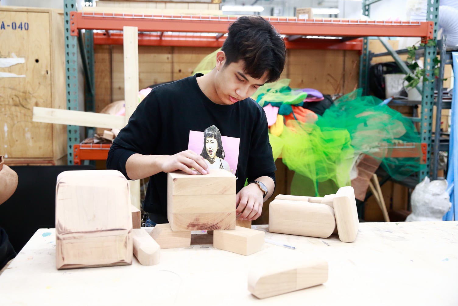 A student at Emily Carr University of Art + Design focuses intently while assembling wooden blocks into a sculptural form. He wears a black long-sleeve shirt with a graphic print and carefully positions a cube atop supporting pieces on a workbench. The background shows a creative studio space with shelving, colourful fabric, and other art materials.