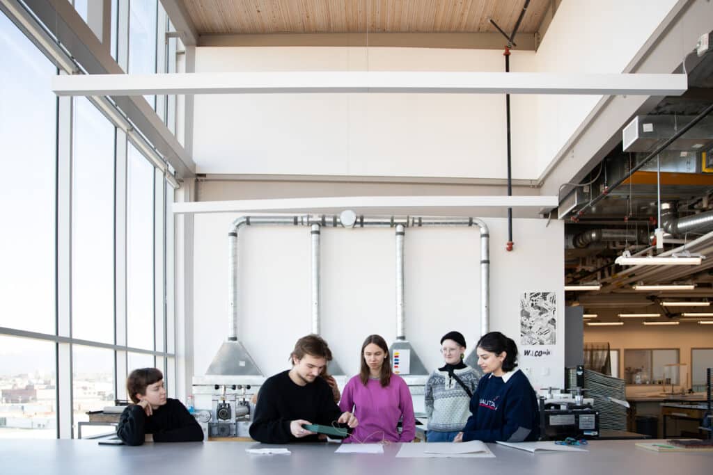 ECU student Eknoor Matharoo collaborates with students from the Estonian Academy of Arts (EKA) during the Tide Places: Artists in Dialogue residency. The group is engaged in a workshop setting, working together at a table with papers and materials spread out. The atmosphere reflects a dynamic exchange of ideas as participants focus on their tasks, showcasing the cross-cultural collaboration facilitated by Canadian artist Lou Sheppard, Estonian artist Laura Põld, and ECU researcher Laura Kozak.