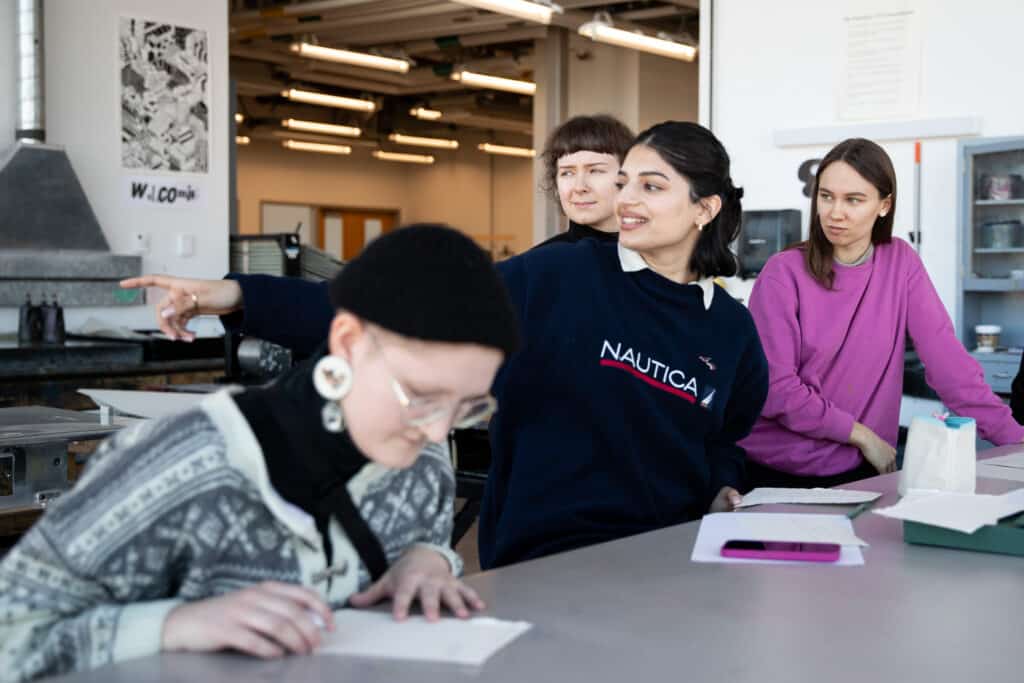 ECU student Eknoor Matharoo collaborates with students from the Estonian Academy of Arts (EKA) during the Tide Places: Artists in Dialogue residency. The group is engaged in a workshop setting, working together at a table with papers and materials spread out. The atmosphere reflects a dynamic exchange of ideas as participants focus on their tasks, showcasing the cross-cultural collaboration facilitated by Canadian artist Lou Sheppard, Estonian artist Laura Põld, and ECU researcher Laura Kozak.