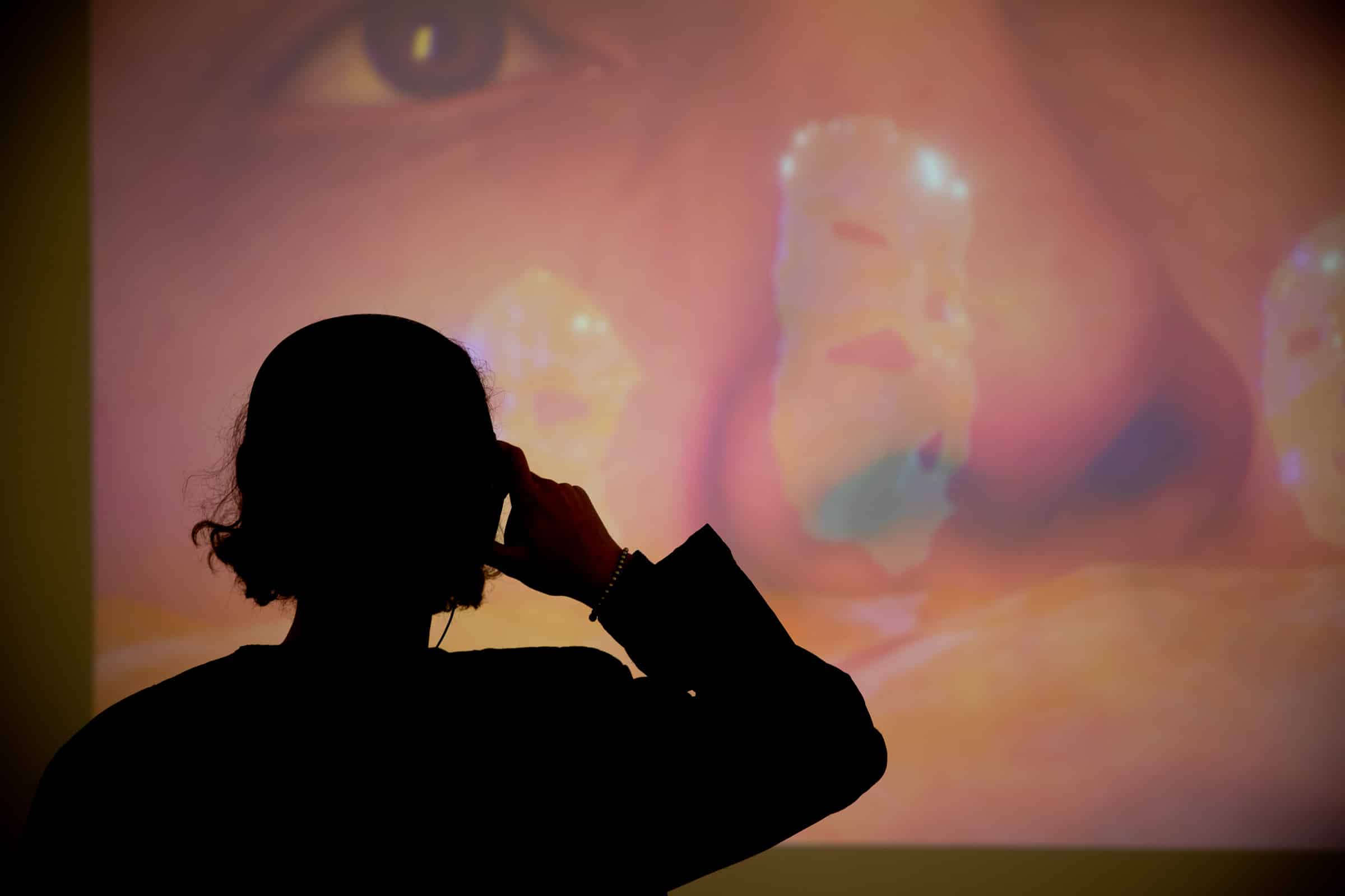A silhouette of a person is captured against a vibrant, colorful projection during the 2022 opening reception for The Show at Emily Carr University. The individual appears to be engaged with the artwork, adjusting their earphones as they immerse themselves in the visual and auditory experience. The warm, dynamic lighting creates a striking contrast, emphasizing the connection between the audience and the art.