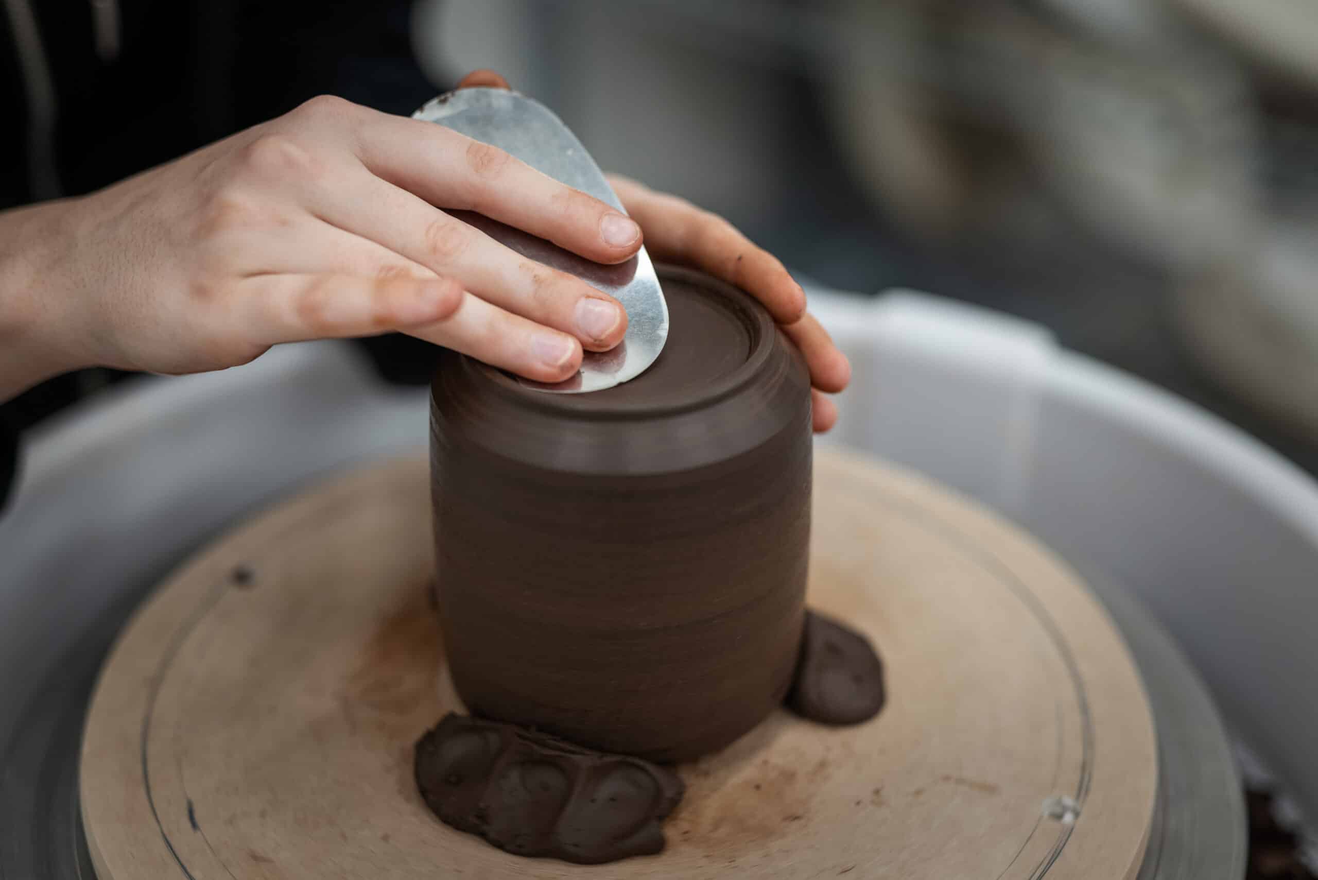 A close-up of hands shaping wet, dark brown clay on a spinning pottery wheel. One hand steadies the clay form while the other uses a metal rib tool to smooth and refine the top surface. The clay is forming a tall, cylindrical vessel, with excess material visible at its base. The background is softly blurred, focusing attention on the tactile process of pottery making.