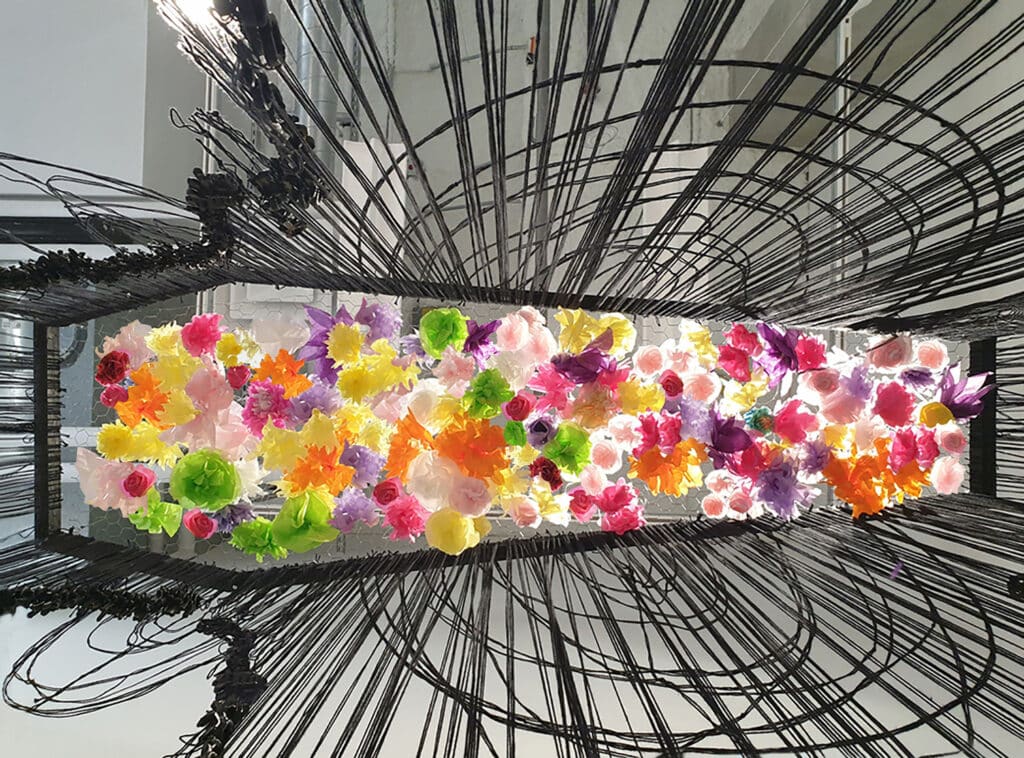 Installation artwork featuring a dense cluster of brightly coloured paper flowers in pink, yellow, orange, green and purple, suspended in the centre of a rectangular frame. Surrounding the flowers are radiating black wires arched in curved patterns, creating a dynamic contrast between the vibrant blooms and the dark structural lines.