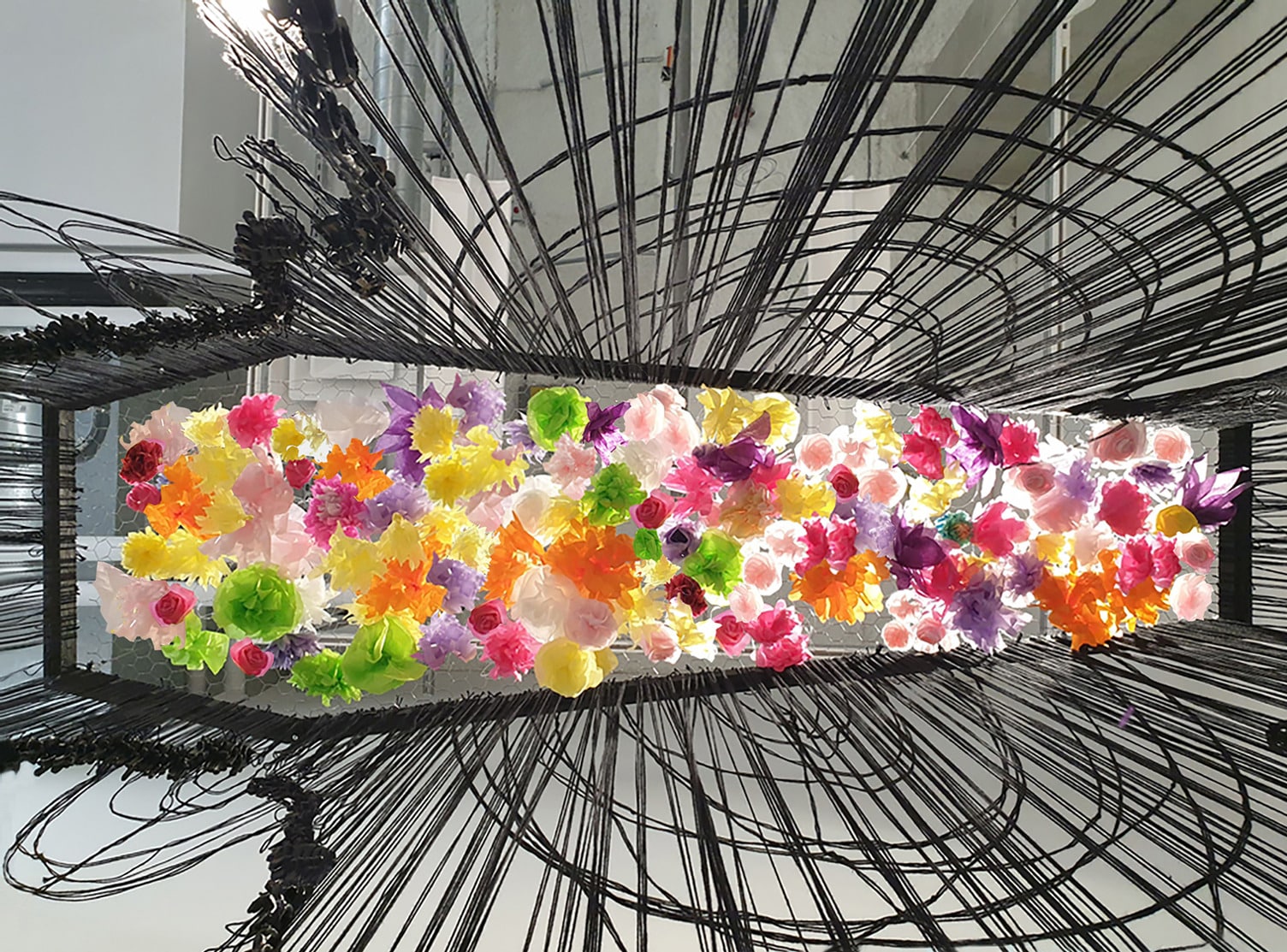 Installation artwork featuring a dense cluster of brightly coloured paper flowers in pink, yellow, orange, green and purple, suspended in the centre of a rectangular frame. Surrounding the flowers are radiating black wires arched in curved patterns, creating a dynamic contrast between the vibrant blooms and the dark structural lines.