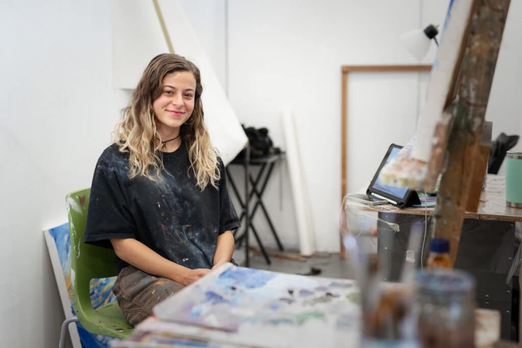 Person with paint splattered black tee-shirt sitting and smiling with painting equipment nearby.