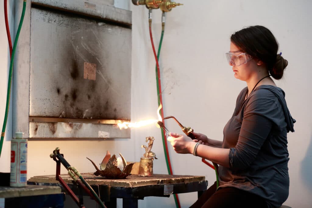 Student wearing safety goggles using a blowtorch to heat metal in a workshop.