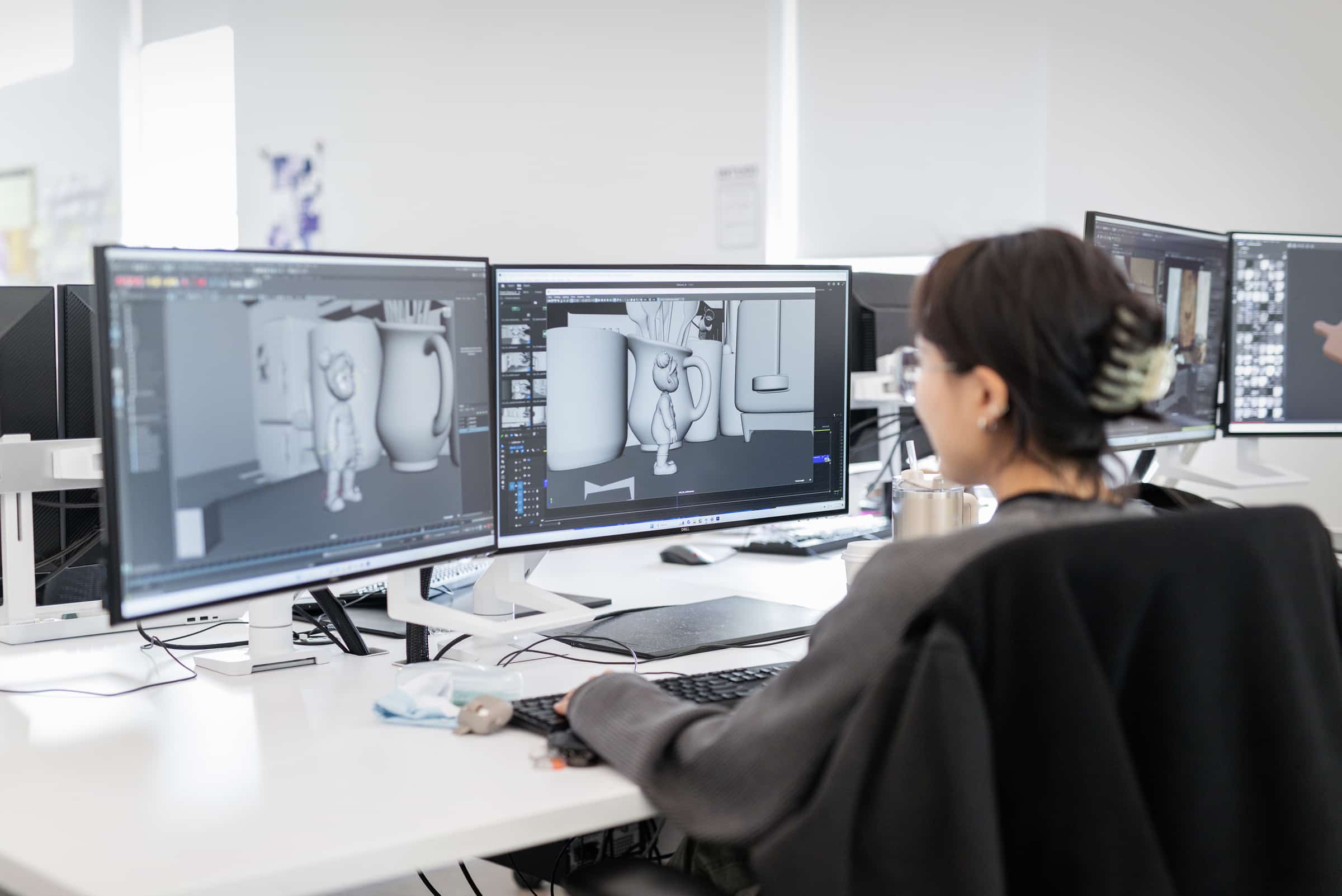 Person working at a desk with multiple monitors, creating a 3D digital scene featuring a small character and oversized objects.