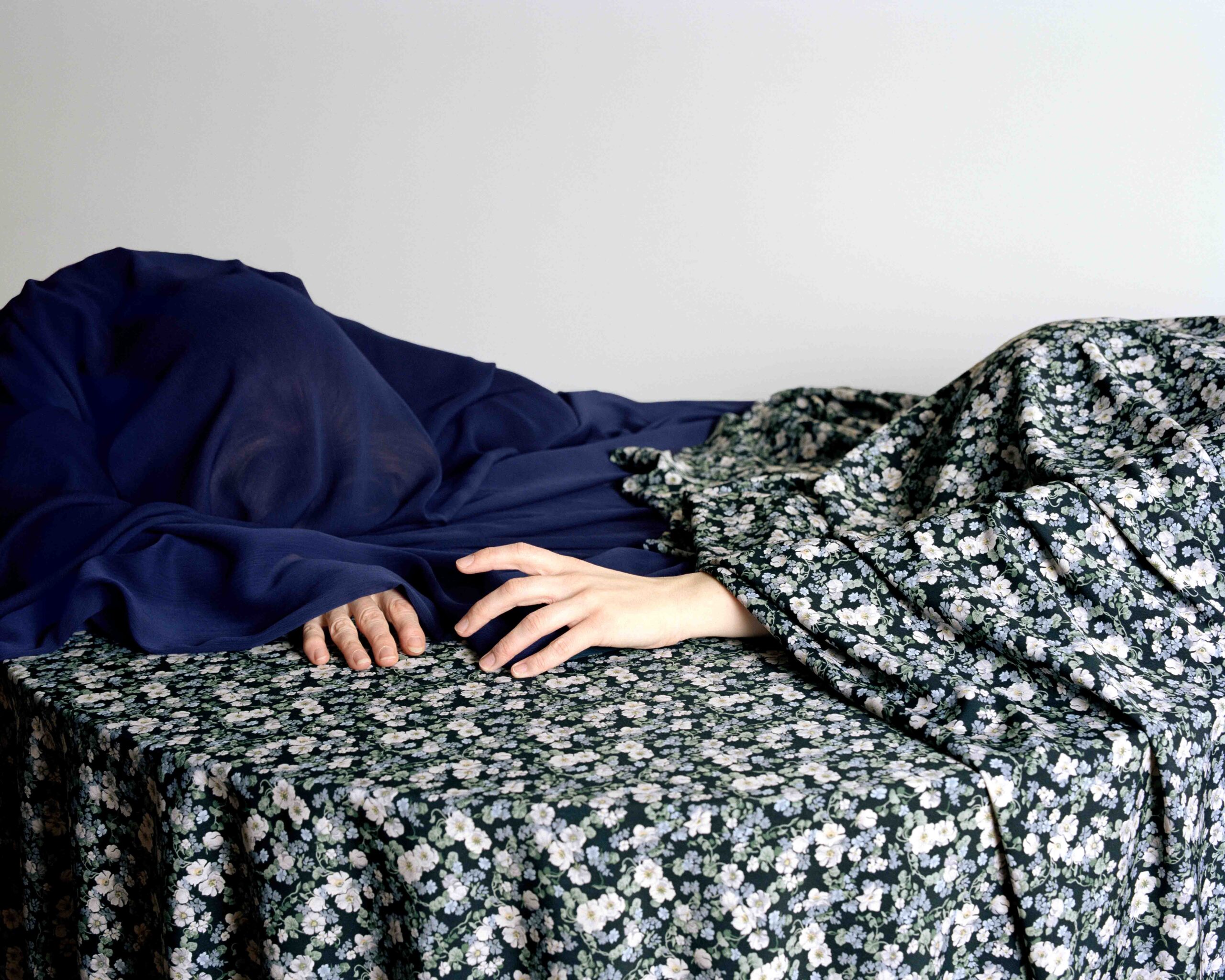 A still-life photograph by Claudia Goulet-Blais (MFA 2025) shows two hands resting gently on a dark green floral-patterned fabric. The body they belong to is concealed beneath layers of draped cloth — one deep navy blue, the other matching the floral sheet. The composition is quiet and intimate, with soft lighting against a plain white wall, emphasizing texture, concealment, and the subtle presence of a hidden figure.