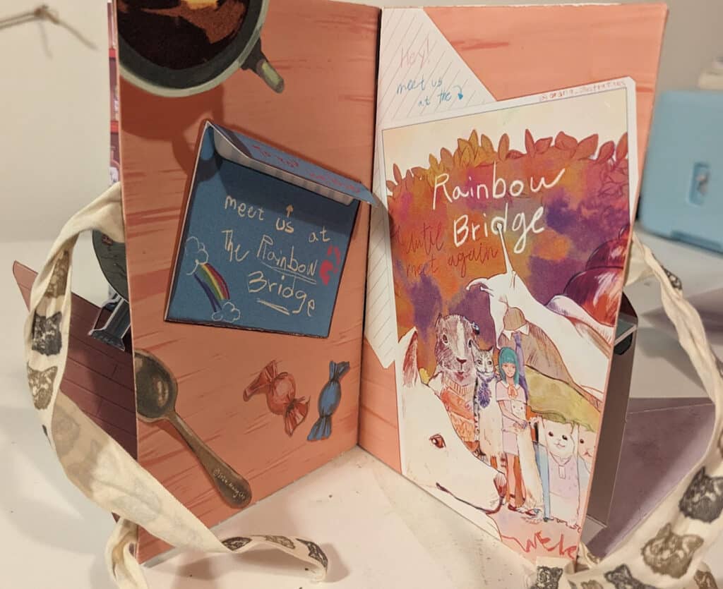 An illustrated pop-up style book spread. On the left, a folded note says “Meet us at The Rainbow Bridge” with small drawings of a rainbow, paw print, candy, and clouds. On the right, a colorful cover illustration shows animals, a girl with blue hair, and text reading “Rainbow Bridge.” The scene is warm and whimsical, tied together with patterned ribbon.
