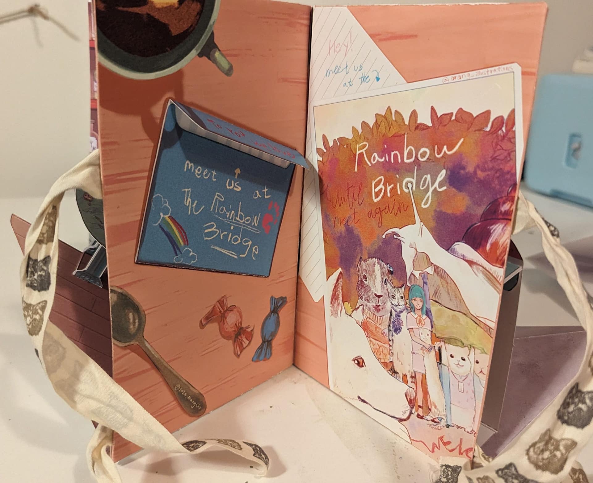 An illustrated pop-up style book spread. On the left, a folded note says “Meet us at The Rainbow Bridge” with small drawings of a rainbow, paw print, candy, and clouds. On the right, a colorful cover illustration shows animals, a girl with blue hair, and text reading “Rainbow Bridge.” The scene is warm and whimsical, tied together with patterned ribbon.