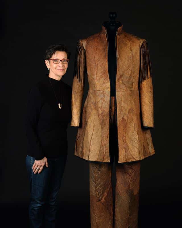 A person stands beside a mannequin wearing a textured brown jacket and pants with fringe details, displayed against a dark background.