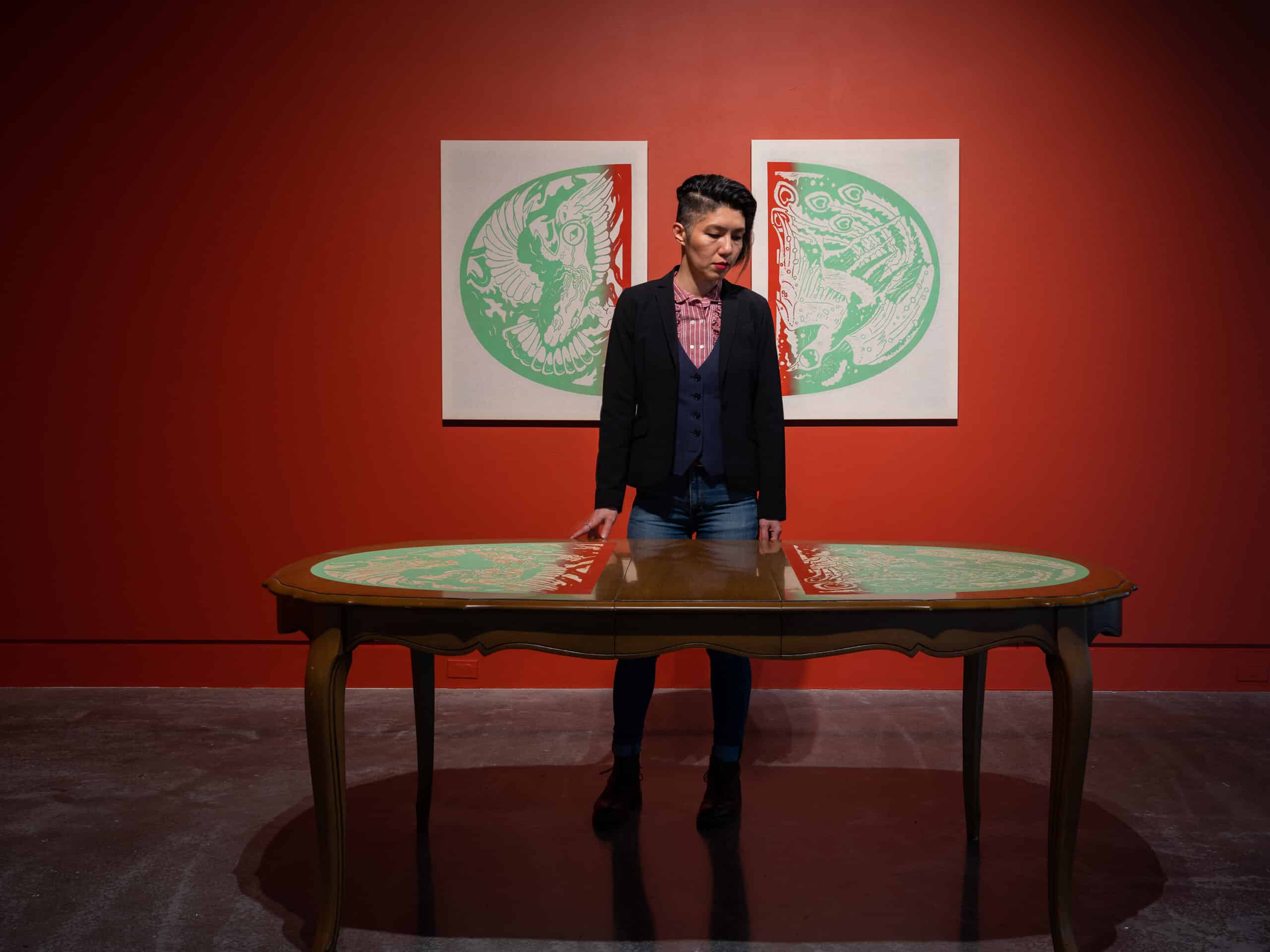 A person stands in a gallery with red walls, looking down at a decorated wooden table, with two green-and-white artworks hanging behind them.
